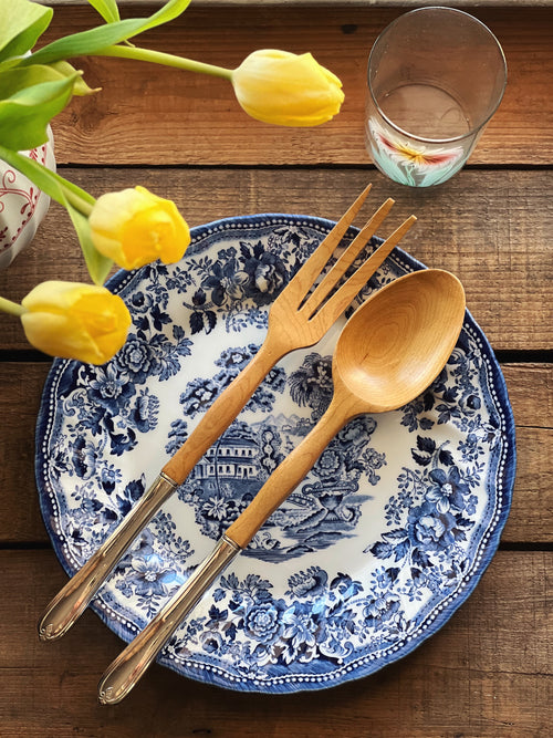 Wooden fork and spoon on a blue and white floral plate with yellow tulips and a glass in the background.