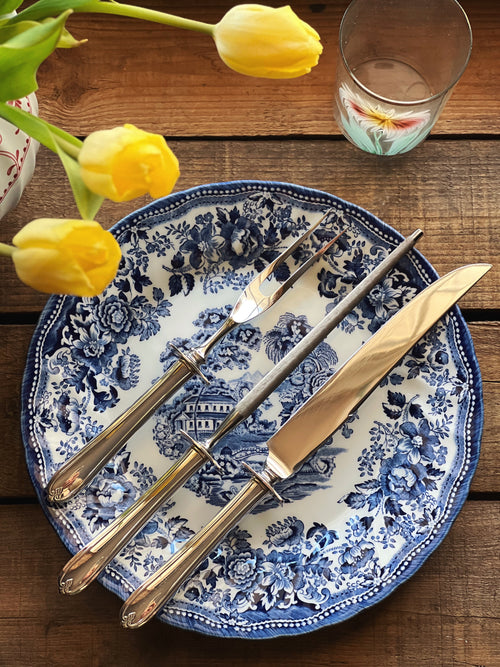 Blue and white floral-patterned plate with silverware on a wooden table with yellow tulips.
