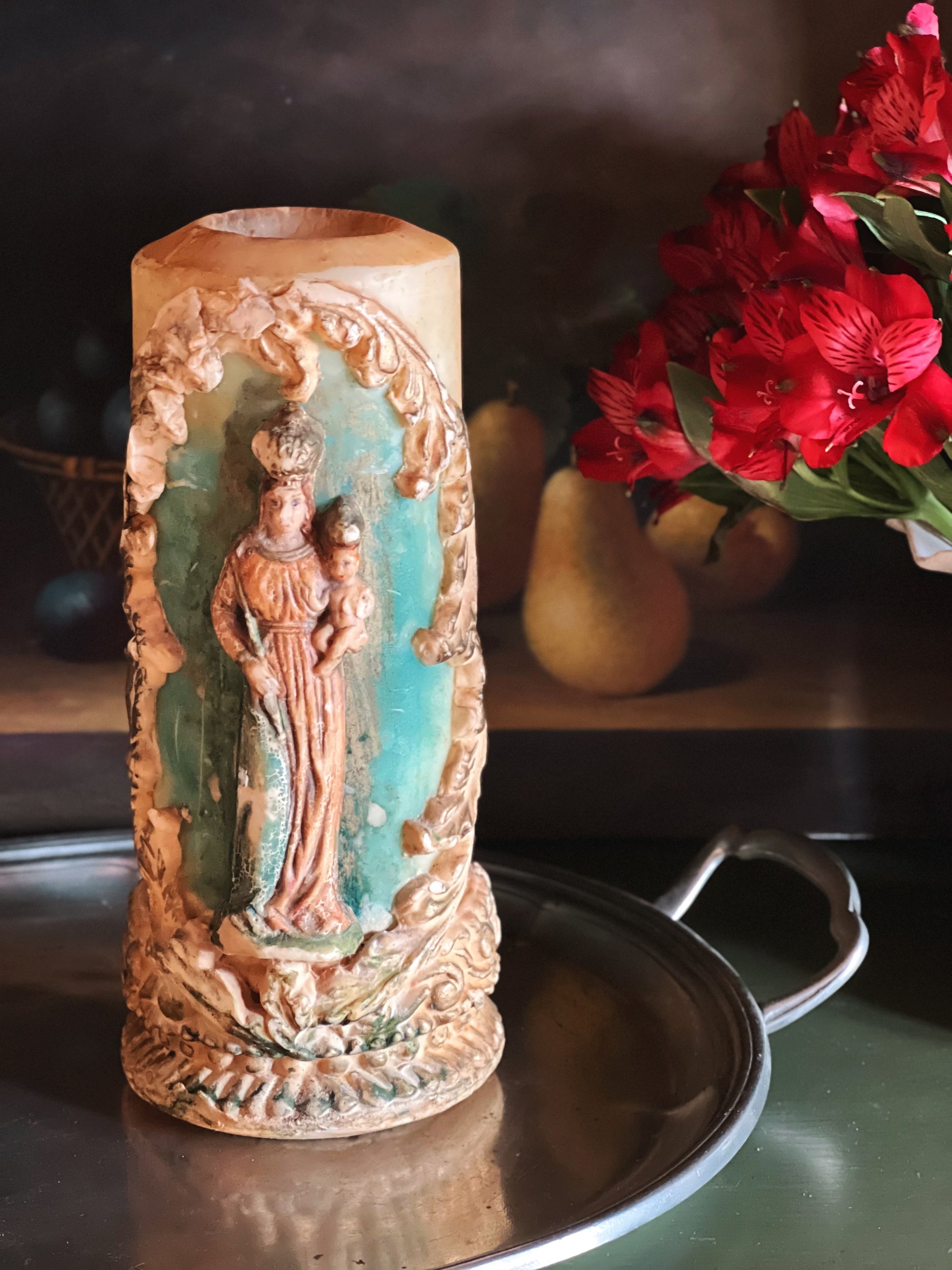 Decorative candle with religious iconography on a reflective surface with flowers in the background