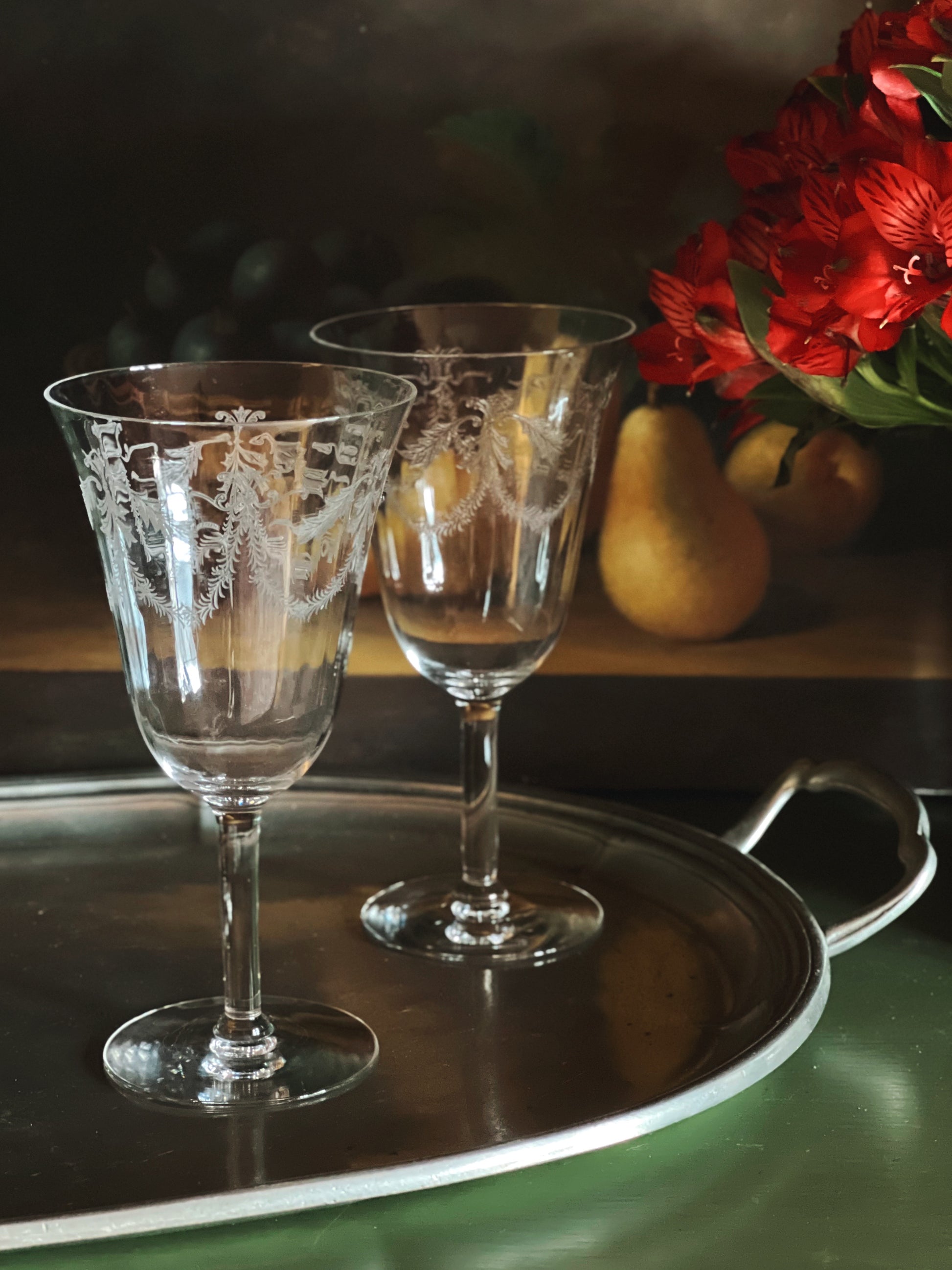 Two etched wine glasses on a metal tray with pears and flowers in the background.