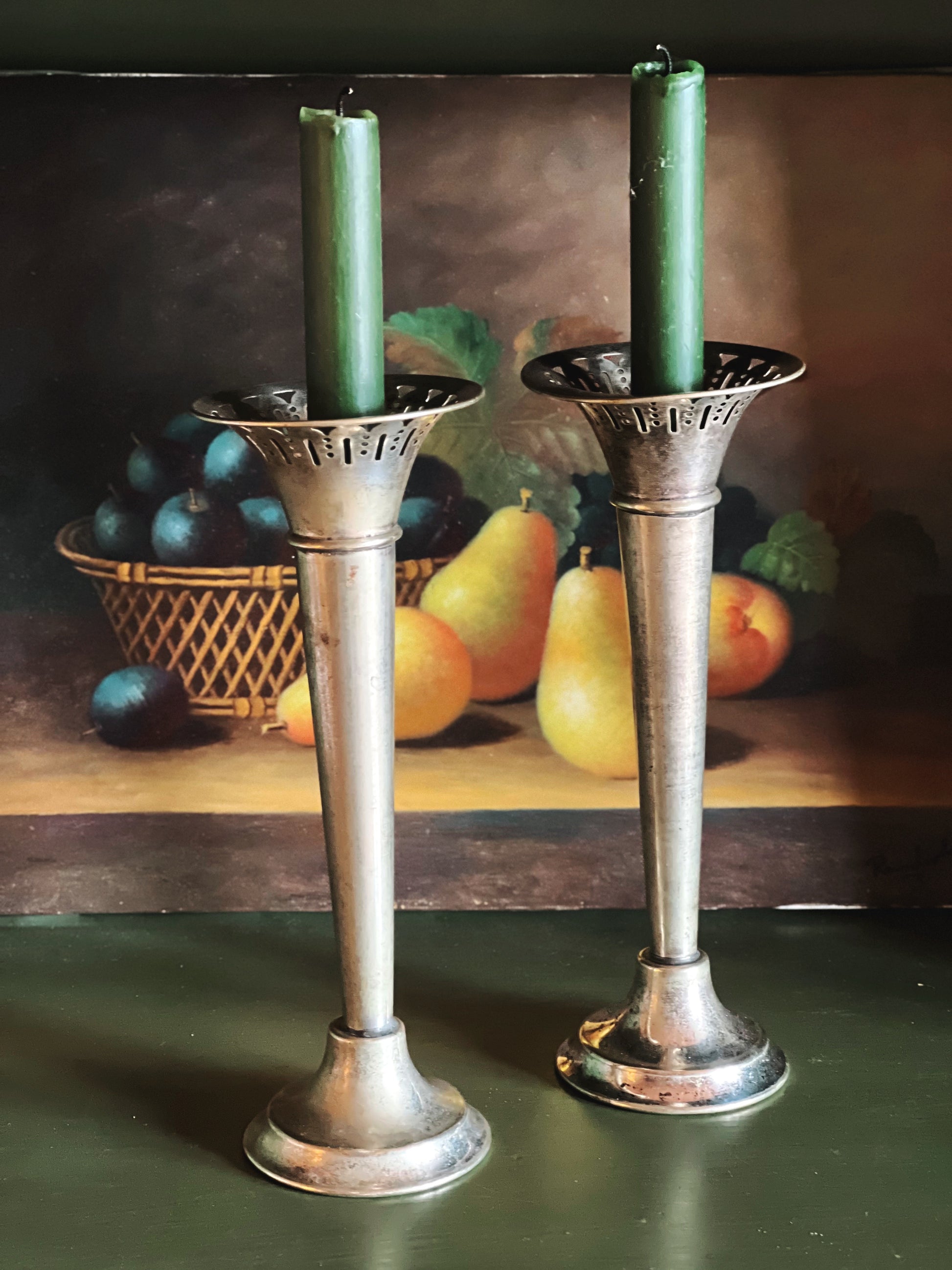 Two silver candle holders with green candles on a table with fruit in the background.
