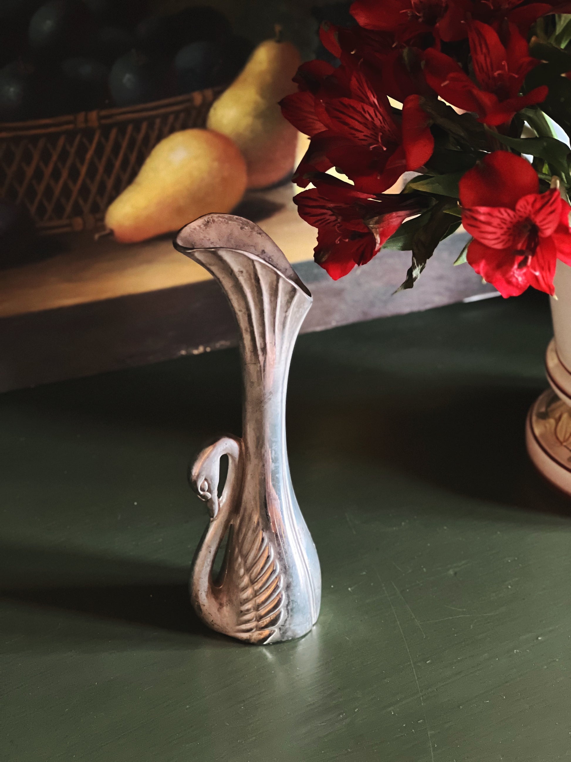Decorative swan vase on a green surface with red flowers and pears in the background