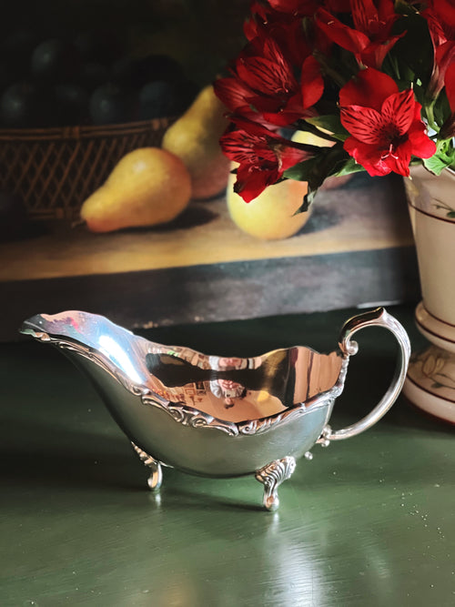 Silver gravy boat on a wooden surface with red flowers and pears in the background