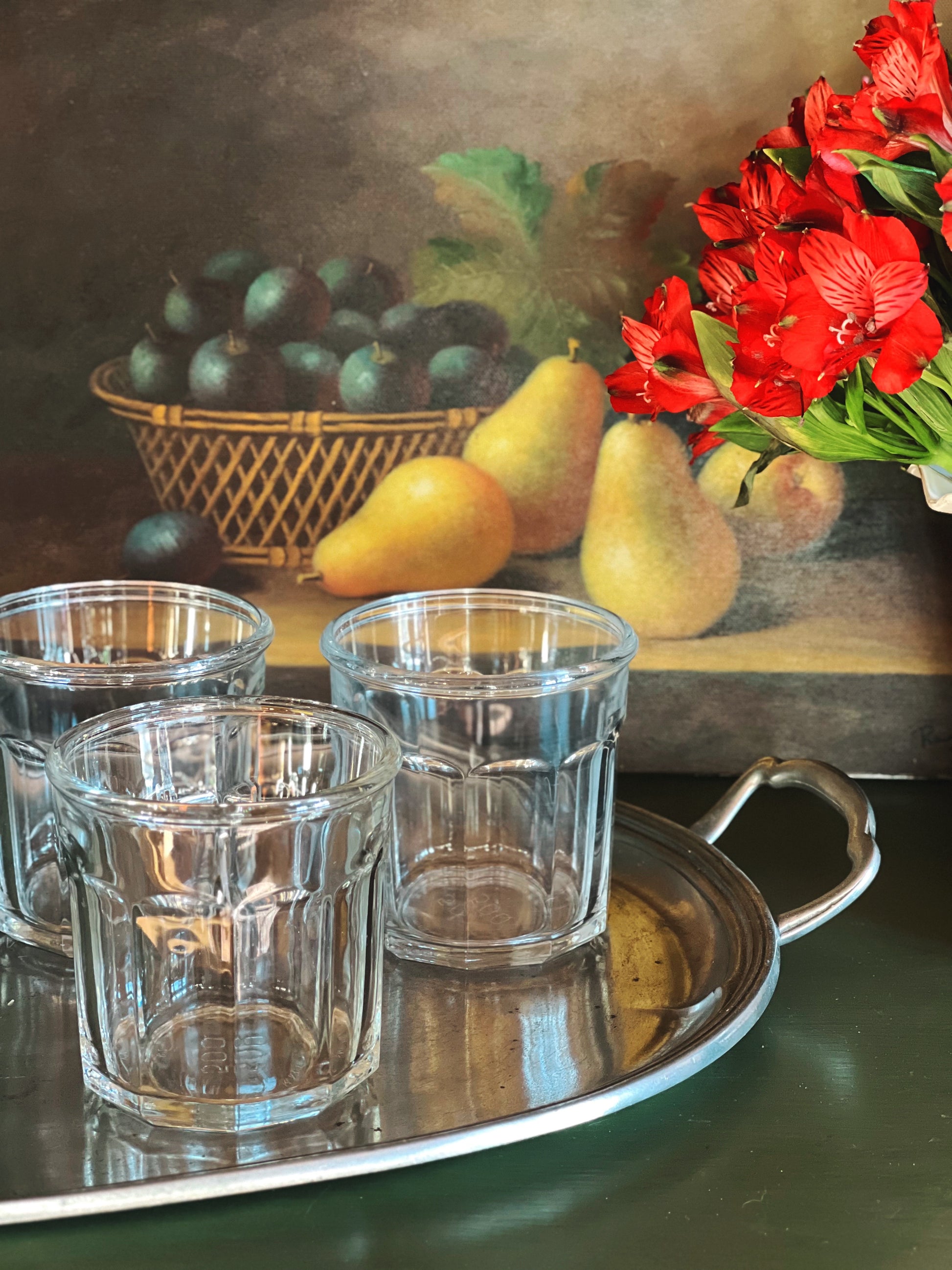 Clear glass tumblers on a silver tray with a still life painting in the background featuring fruits and flowers.
