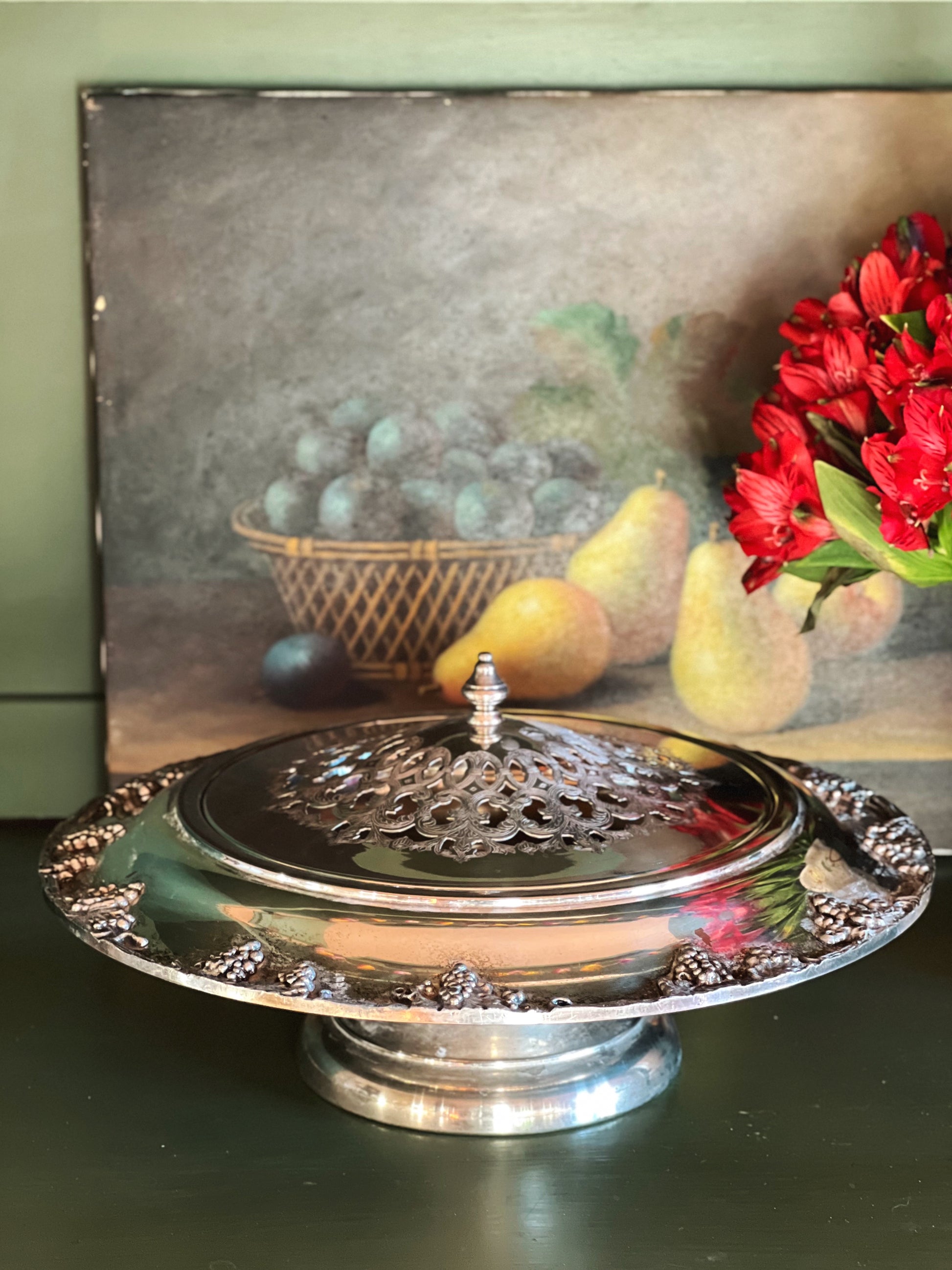 Silver cake stand with decorative lid in front of a still life painting
