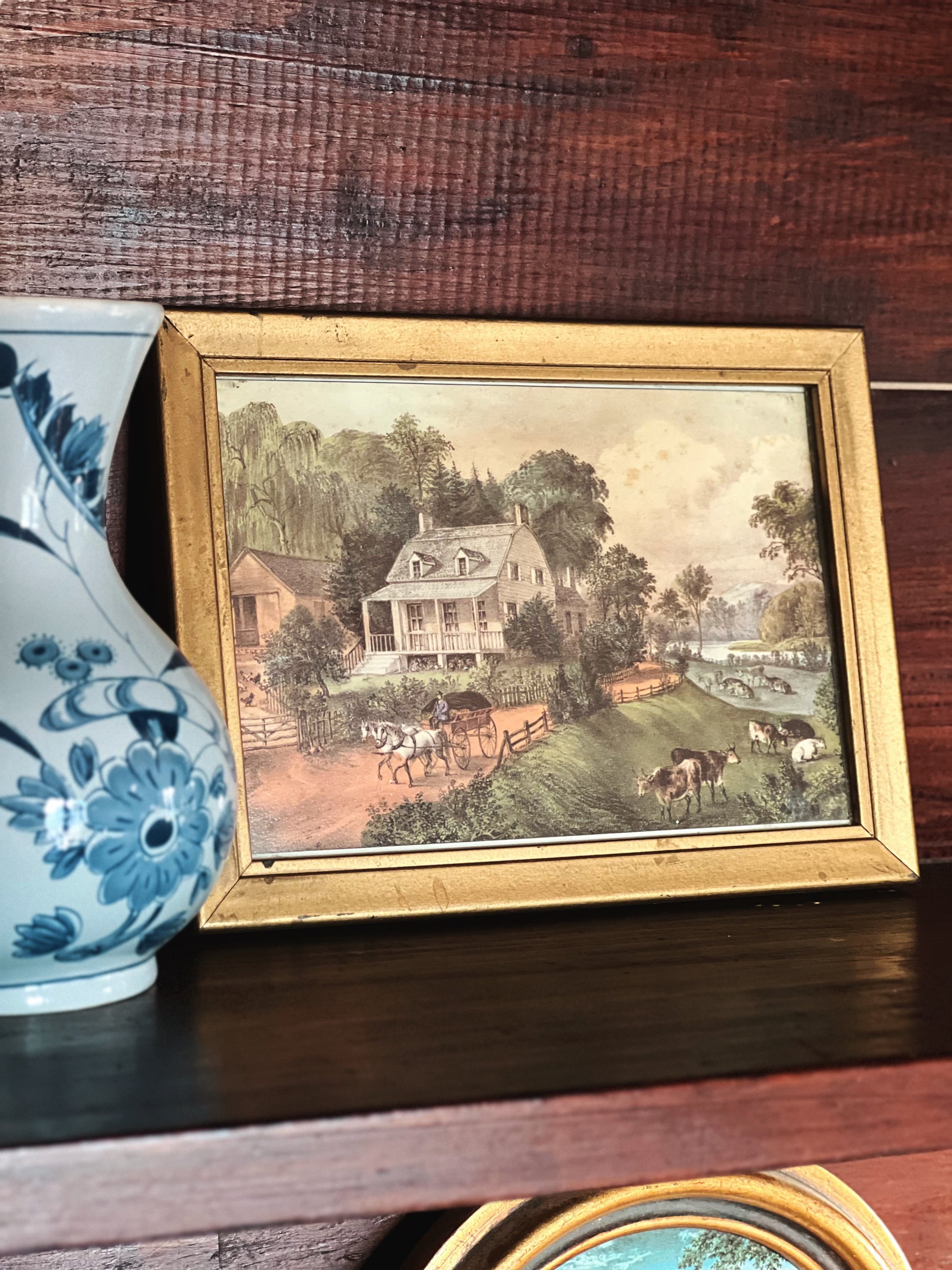 Framed artwork of a pastoral scene with a house and animals on a wooden surface.