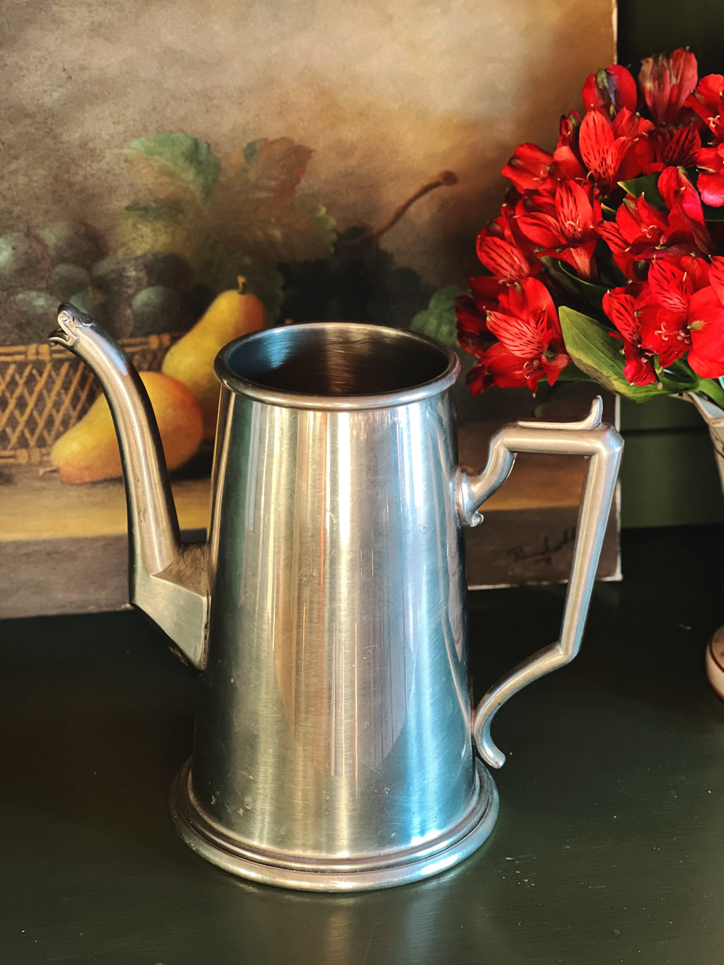 Silver coffee pot on a dark surface with a painting and flowers in the background