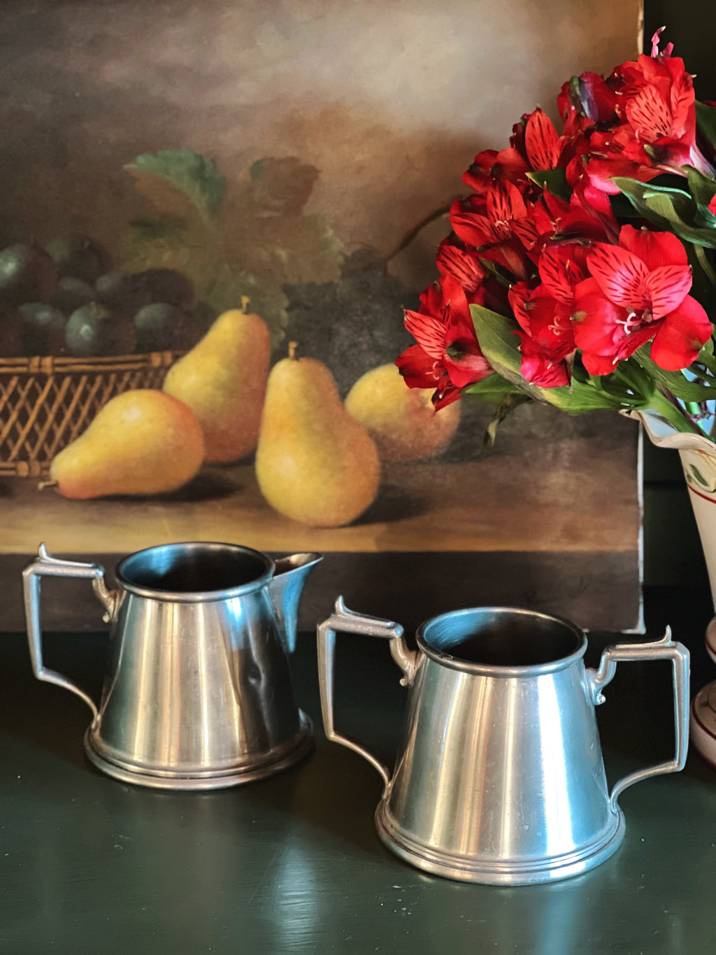 Two metal mugs on a table with a still life painting of pears and flowers in the background.