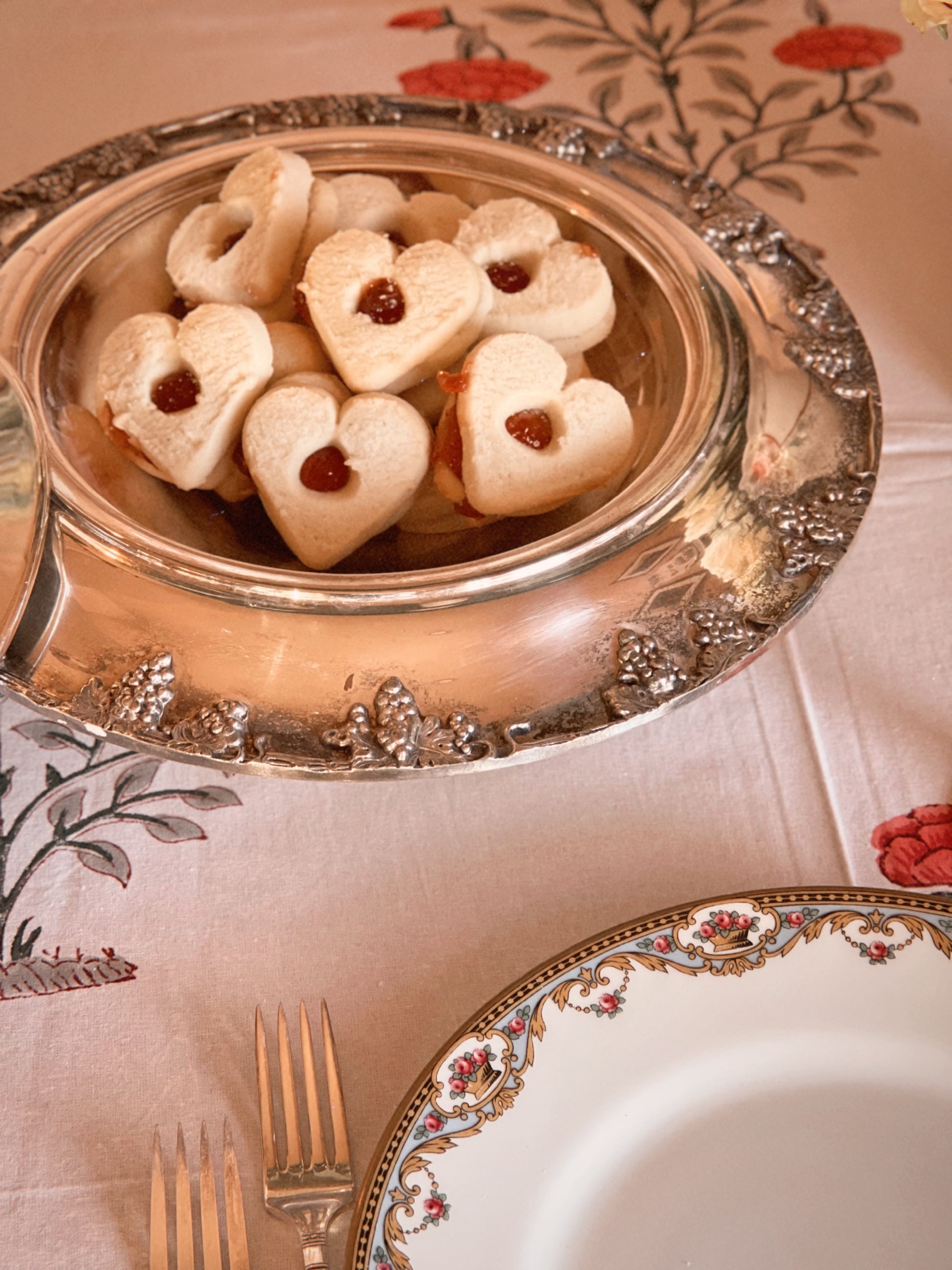 Heart-shaped cookies with jam filling on a decorative silver platter on a patterned tablecloth.