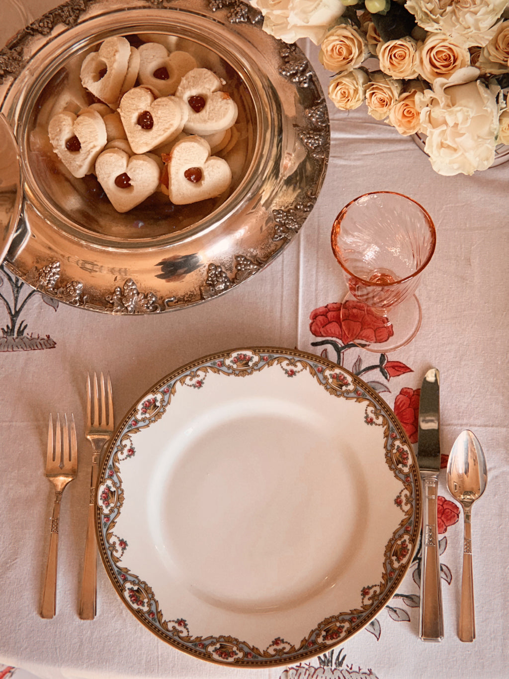 Dinner setting with decorative plates, silverware, and a bowl of heart-shaped cookies on a tablecloth.