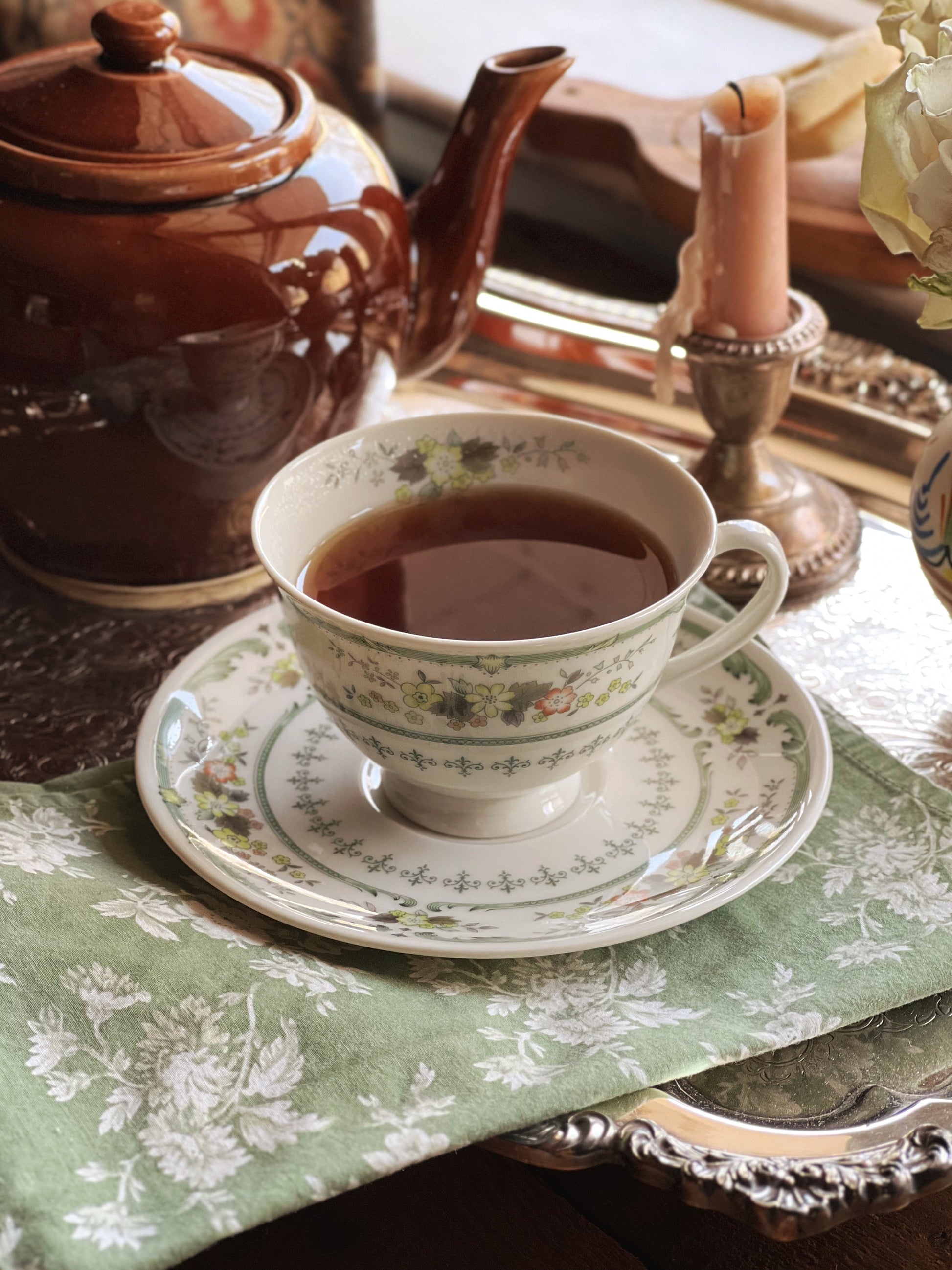 Tea cup with floral design on a saucer, next to a brown teapot on a patterned tablecloth.