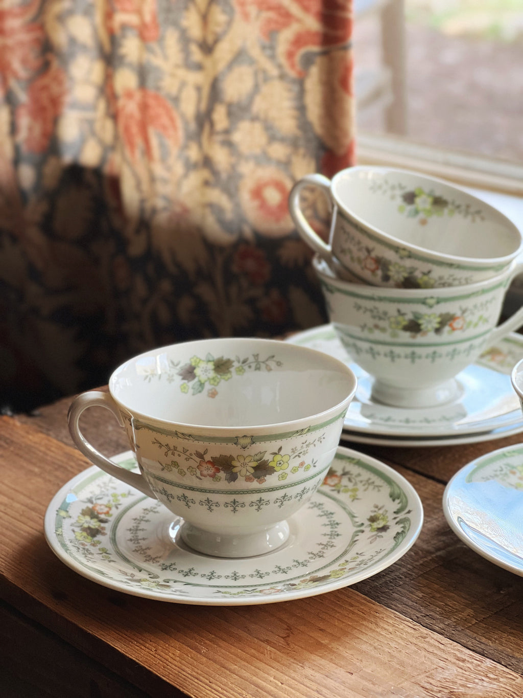 Set of floral teacups and saucers on a wooden table with a patterned curtain in the background.