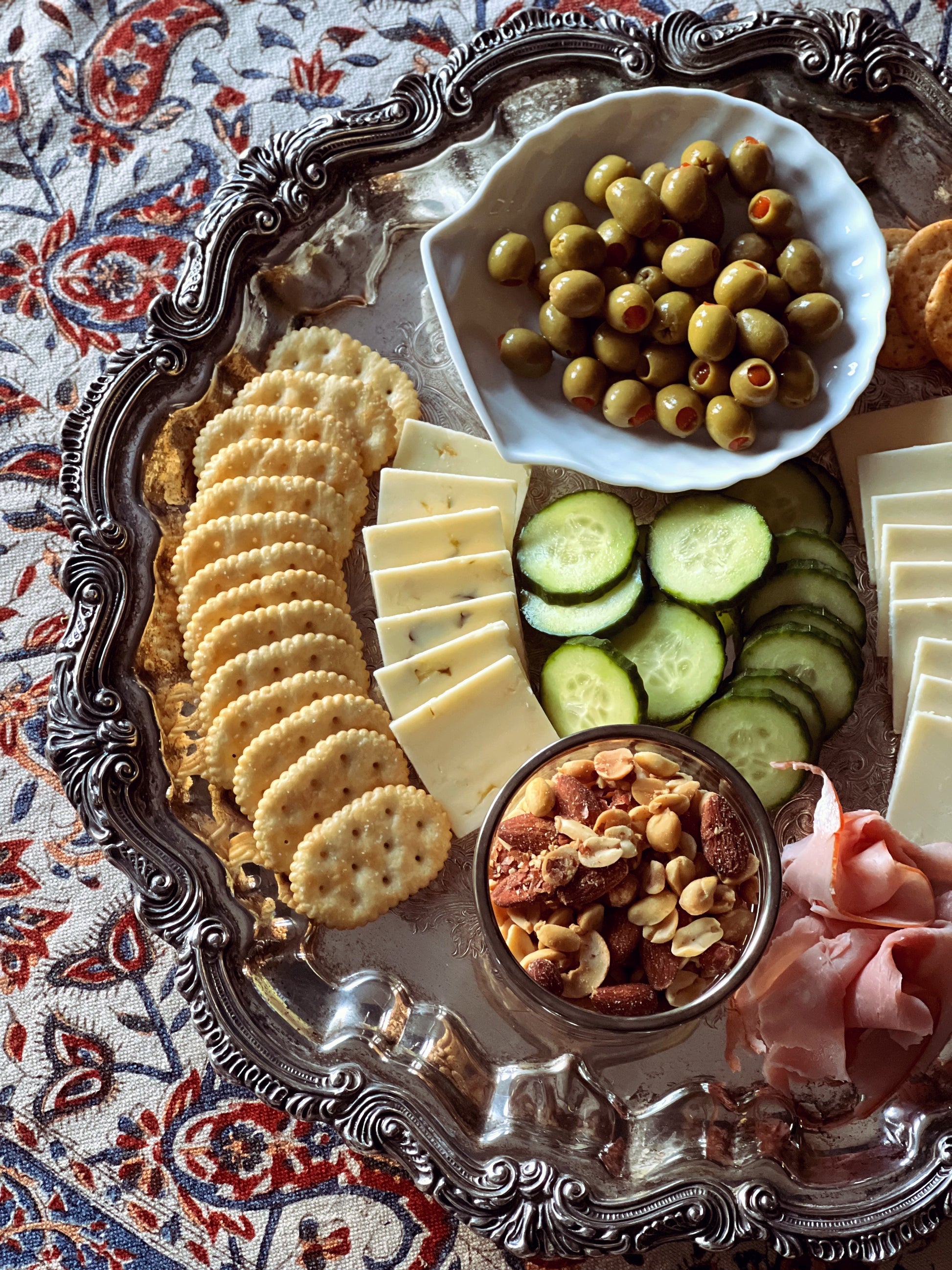 Platter with assorted snacks including crackers, cheese, olives, and dried fruits on a patterned tablecloth.