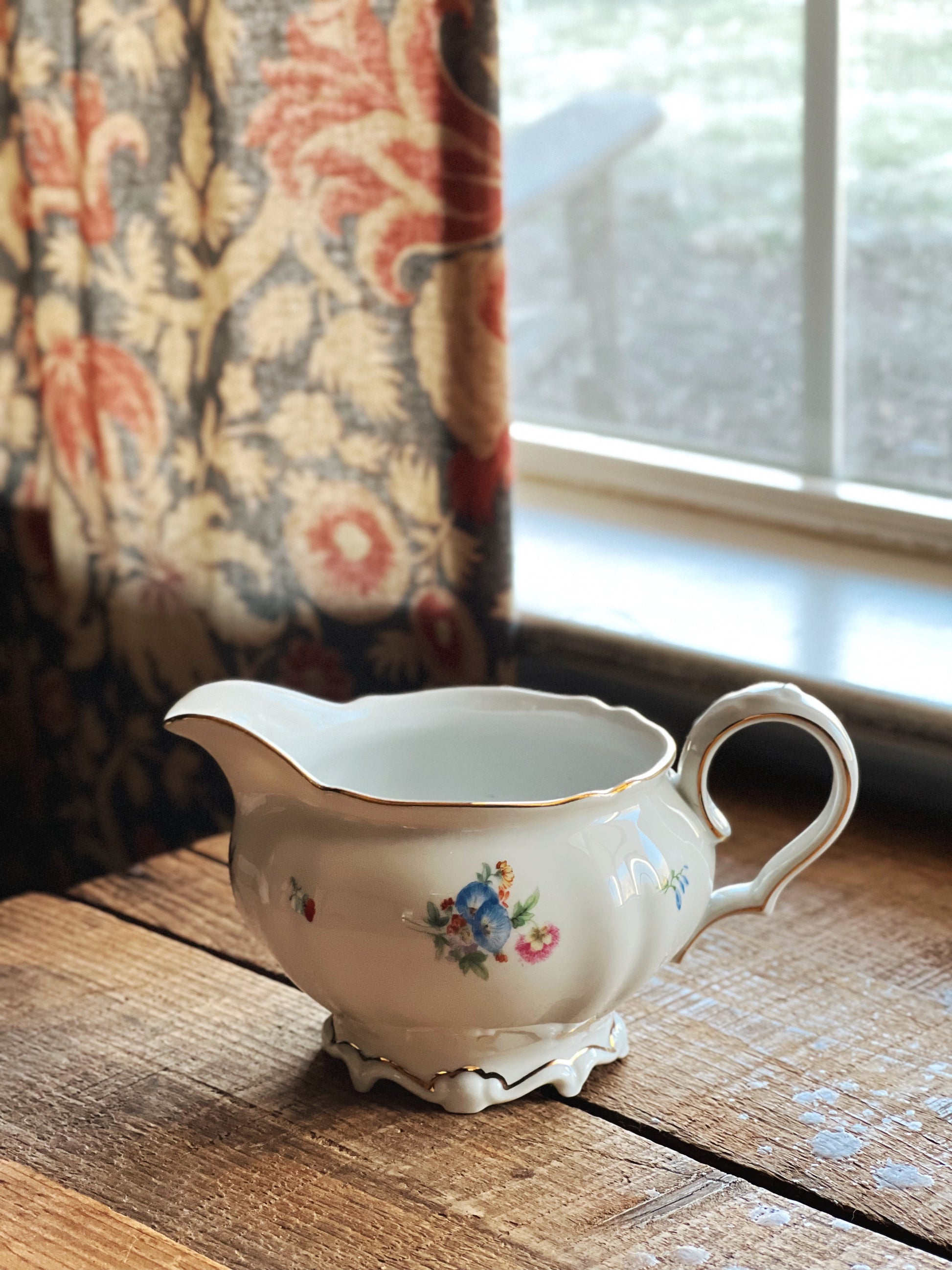 Decorative ceramic pitcher on a wooden surface with a window and curtain in the background