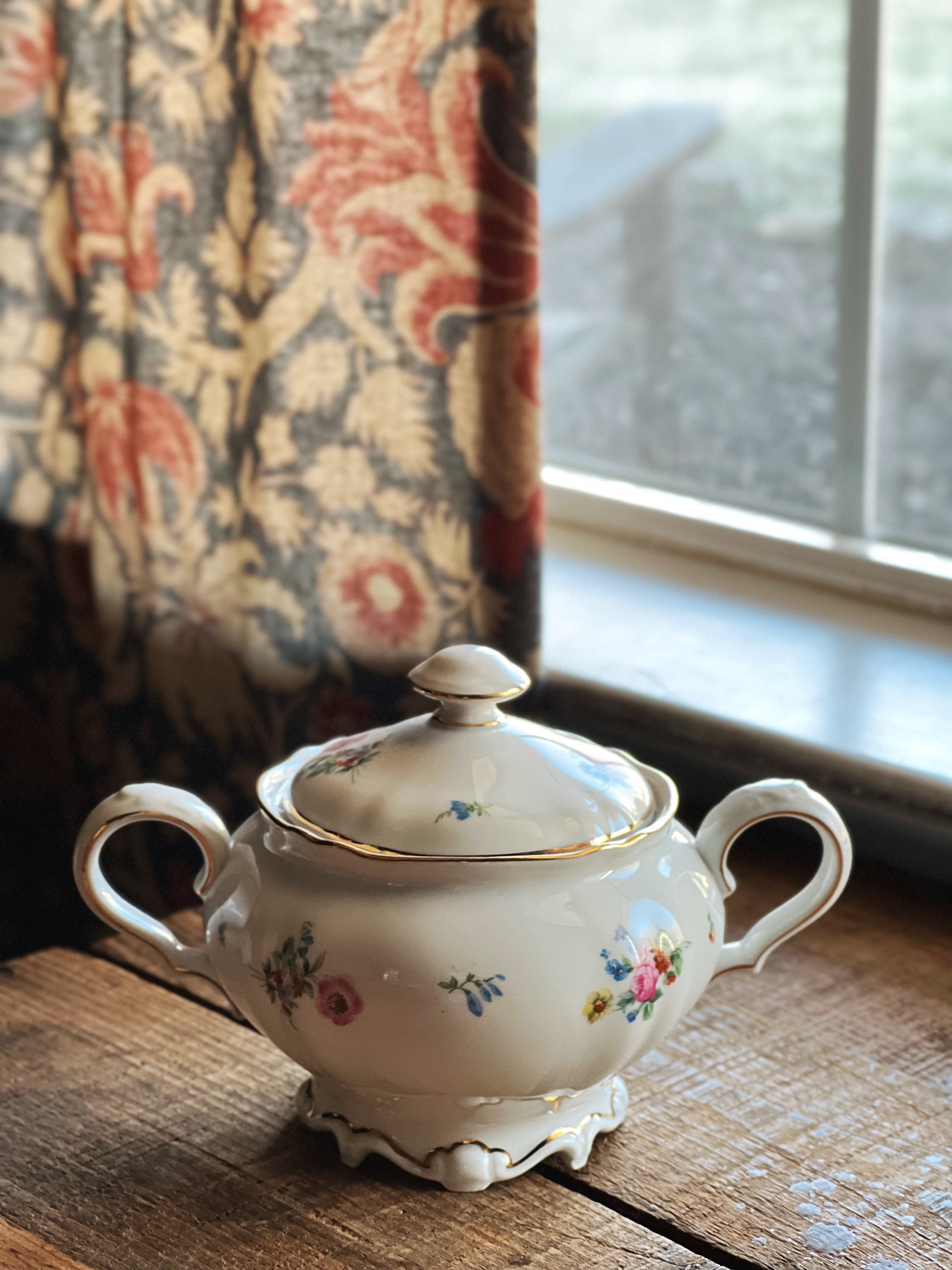 Decorative ceramic sugar bowl with floral patterns on a wooden surface near a window.