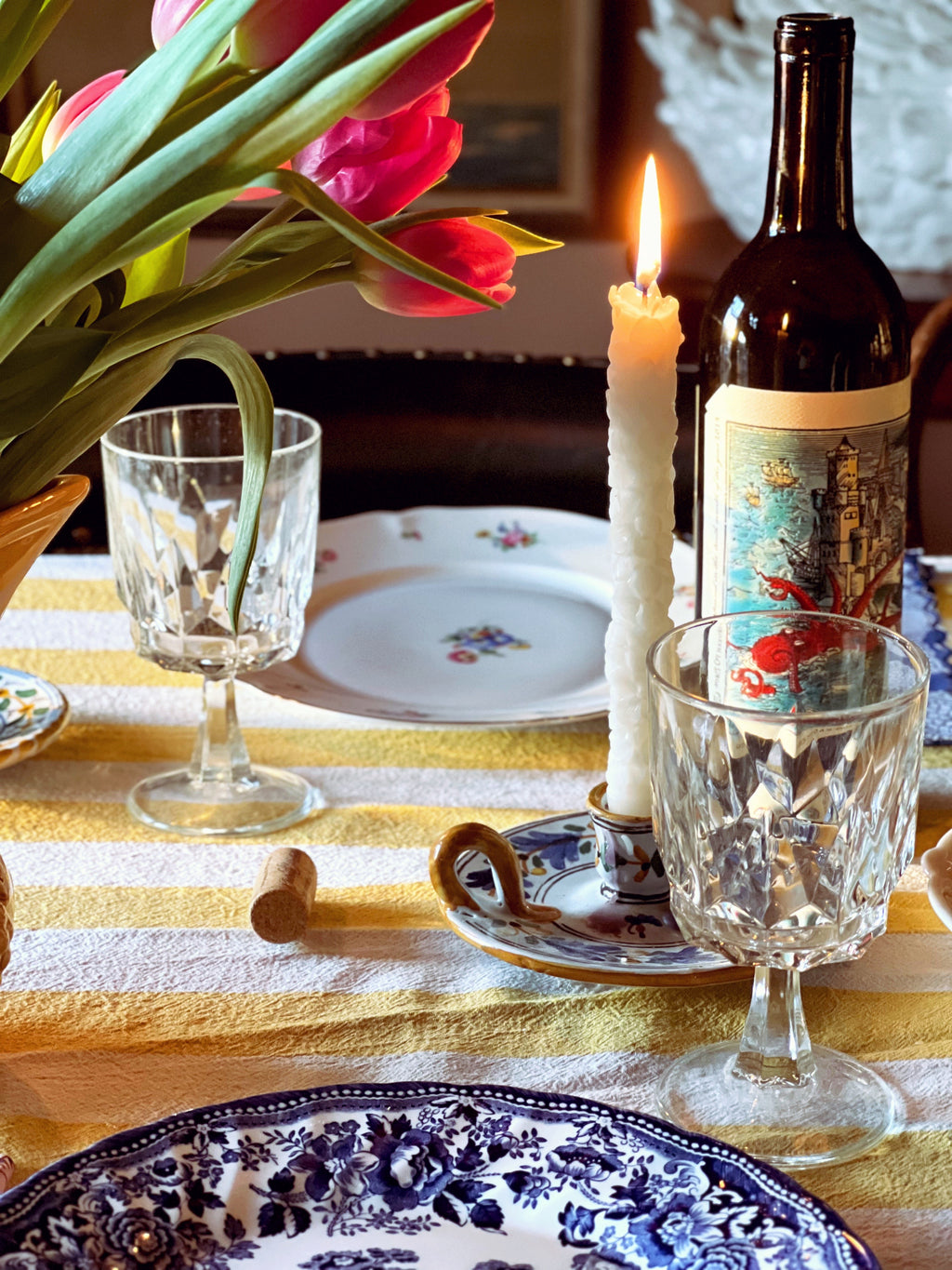 Dining table setting with tulips, wine bottle, glasses, and candles on a striped tablecloth.