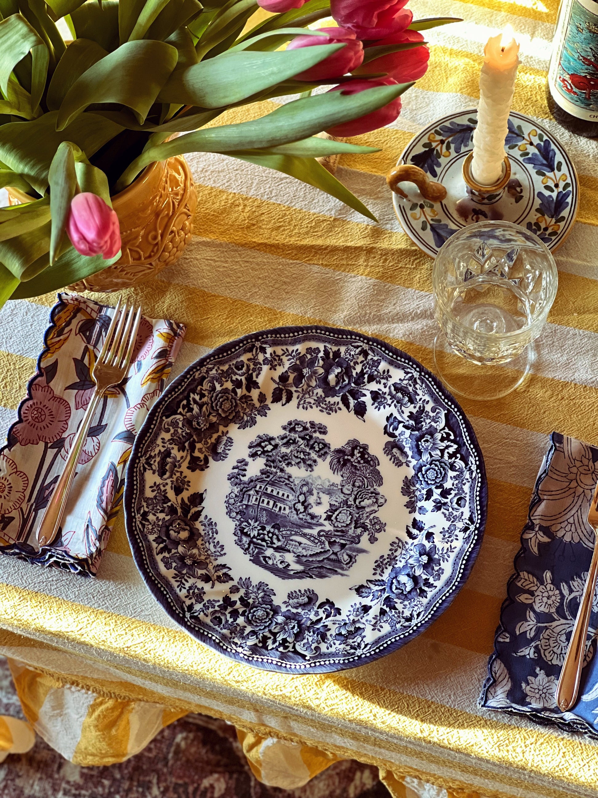 Decorative table setting with a blue and white plate, floral arrangement, and candles on a yellow tablecloth.