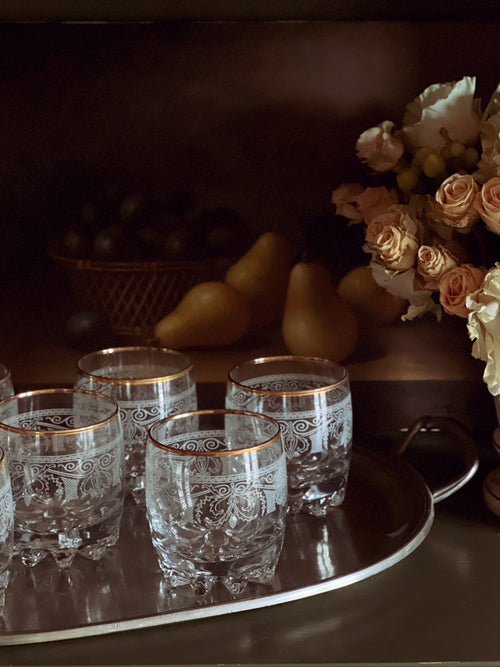 Set of four ornate glasses on a tray with a blurred background of fruits and flowers.