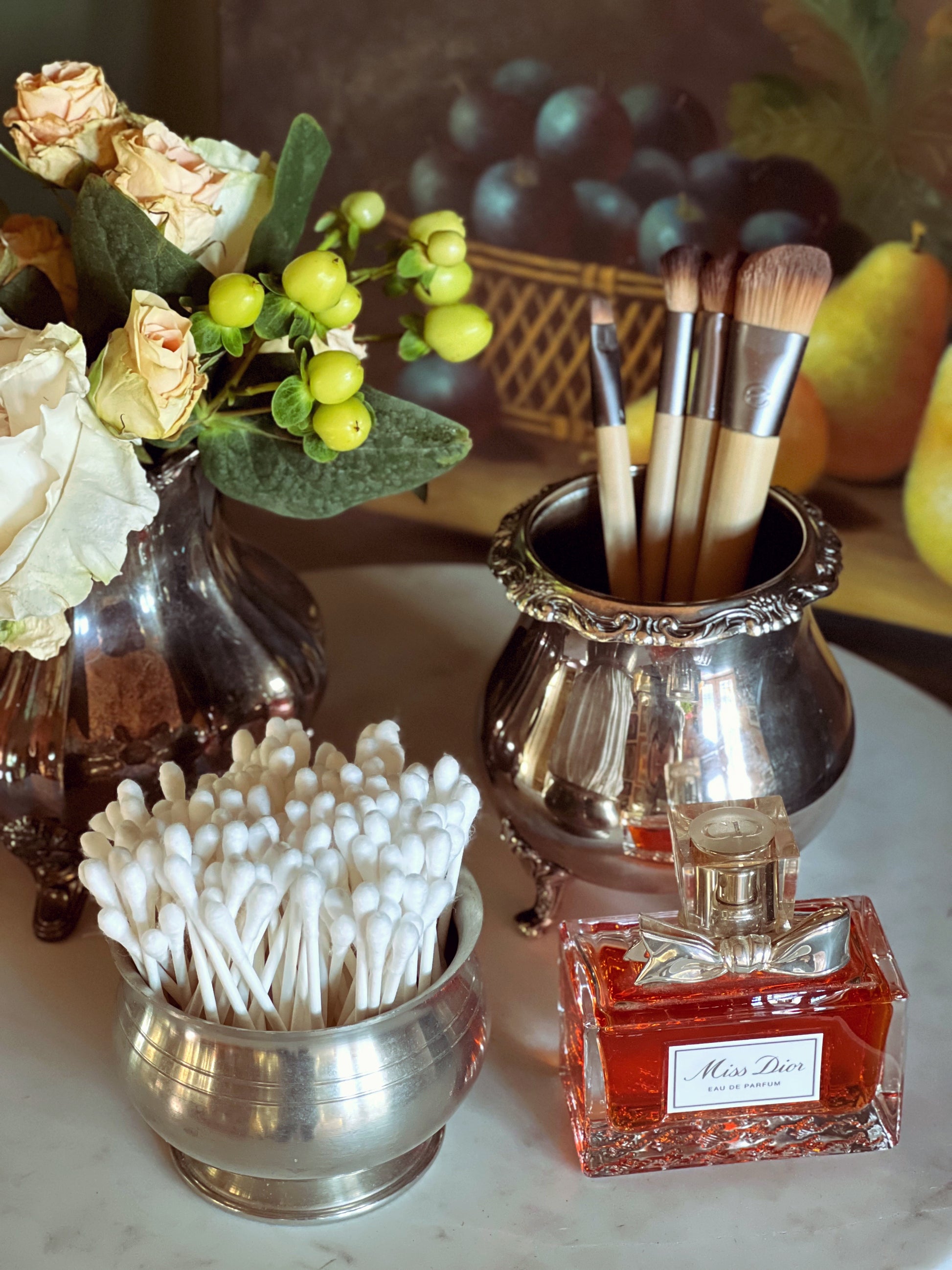 Silver containers with cotton swabs, makeup brushes, and a perfume bottle on a marble surface.