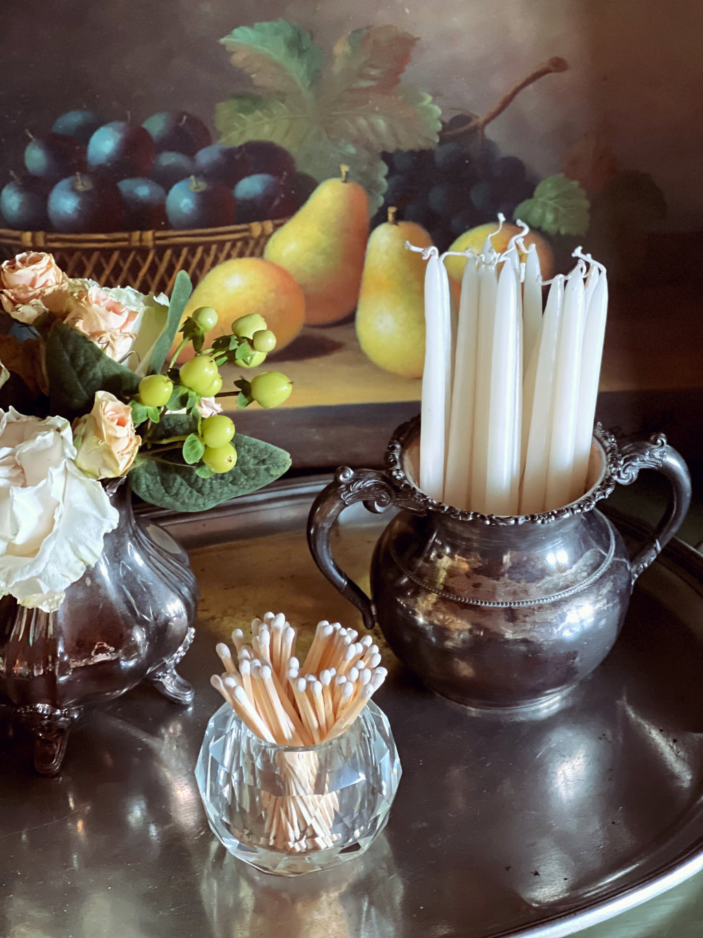 Still life arrangement with candles, fruits, and flowers on a reflective surface.