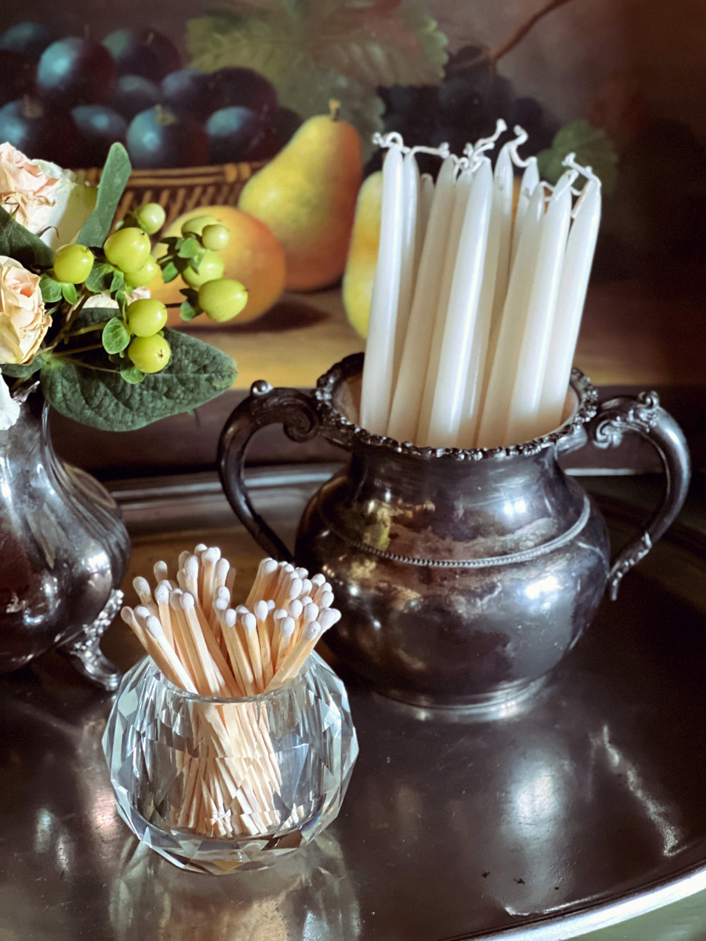 Two containers of toothpicks on a reflective surface with fruit and flowers in the background.