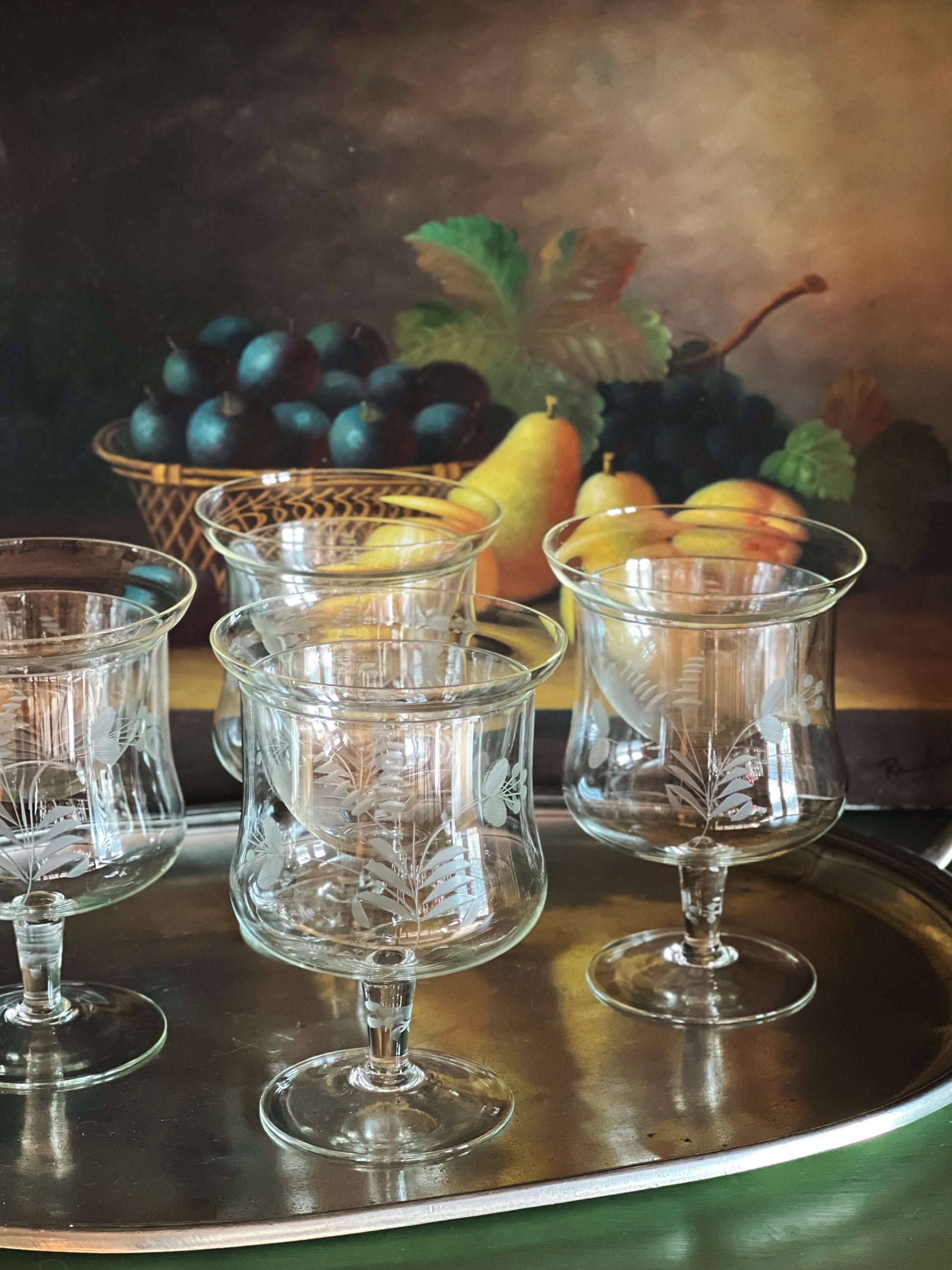 Set of glass goblets on a reflective surface with a still life background featuring fruits.