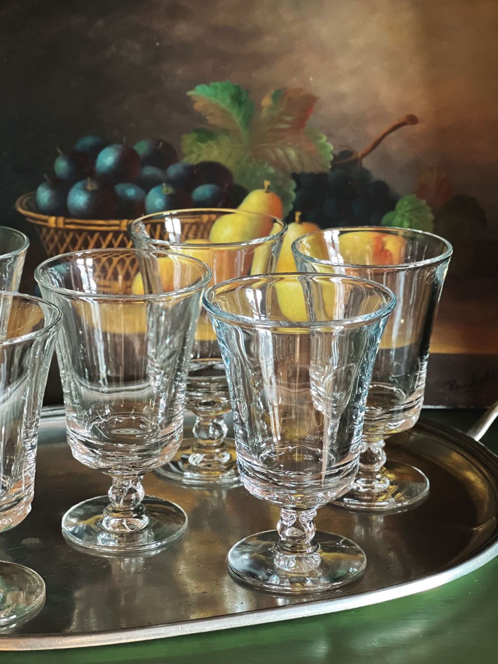 Set of clear glass goblets on a tray with a still life painting in the background