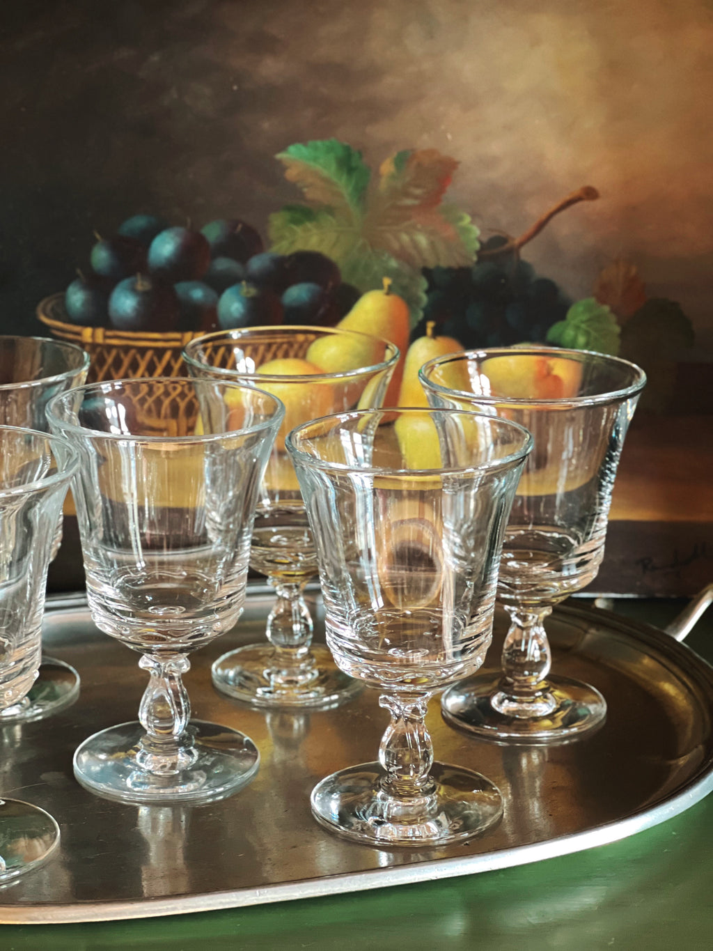 Set of clear glass goblets on a silver tray with a still life background of fruits.
