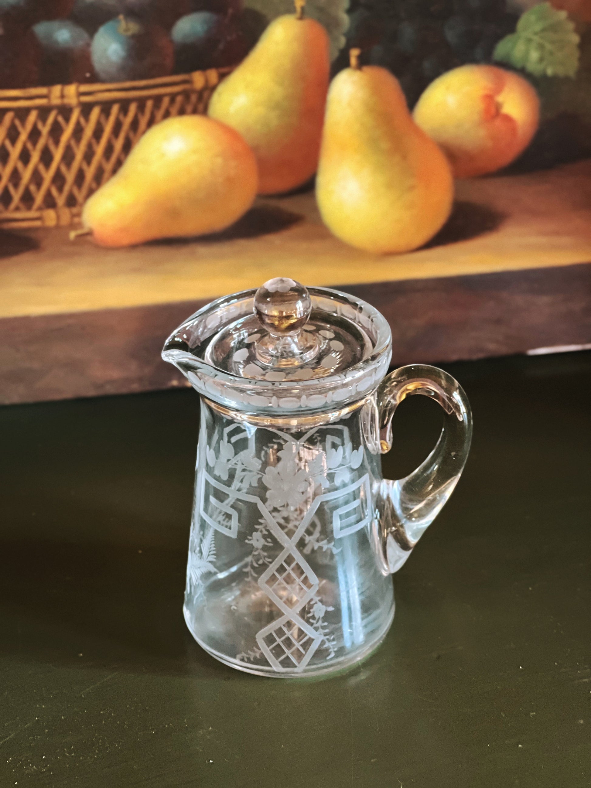 Clear glass pitcher with intricate designs on a dark surface, with pears in the background.