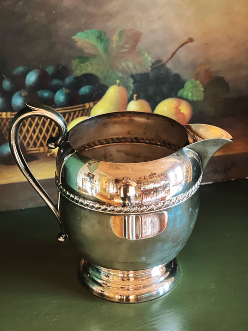 Silver pitcher on a green surface with a fruit painting in the background