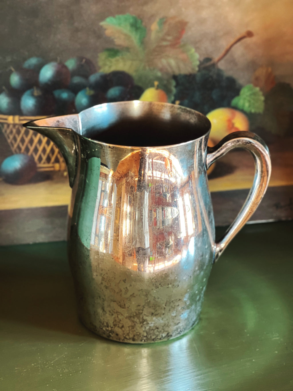 Silver pitcher on a reflective surface with a fruit painting in the background