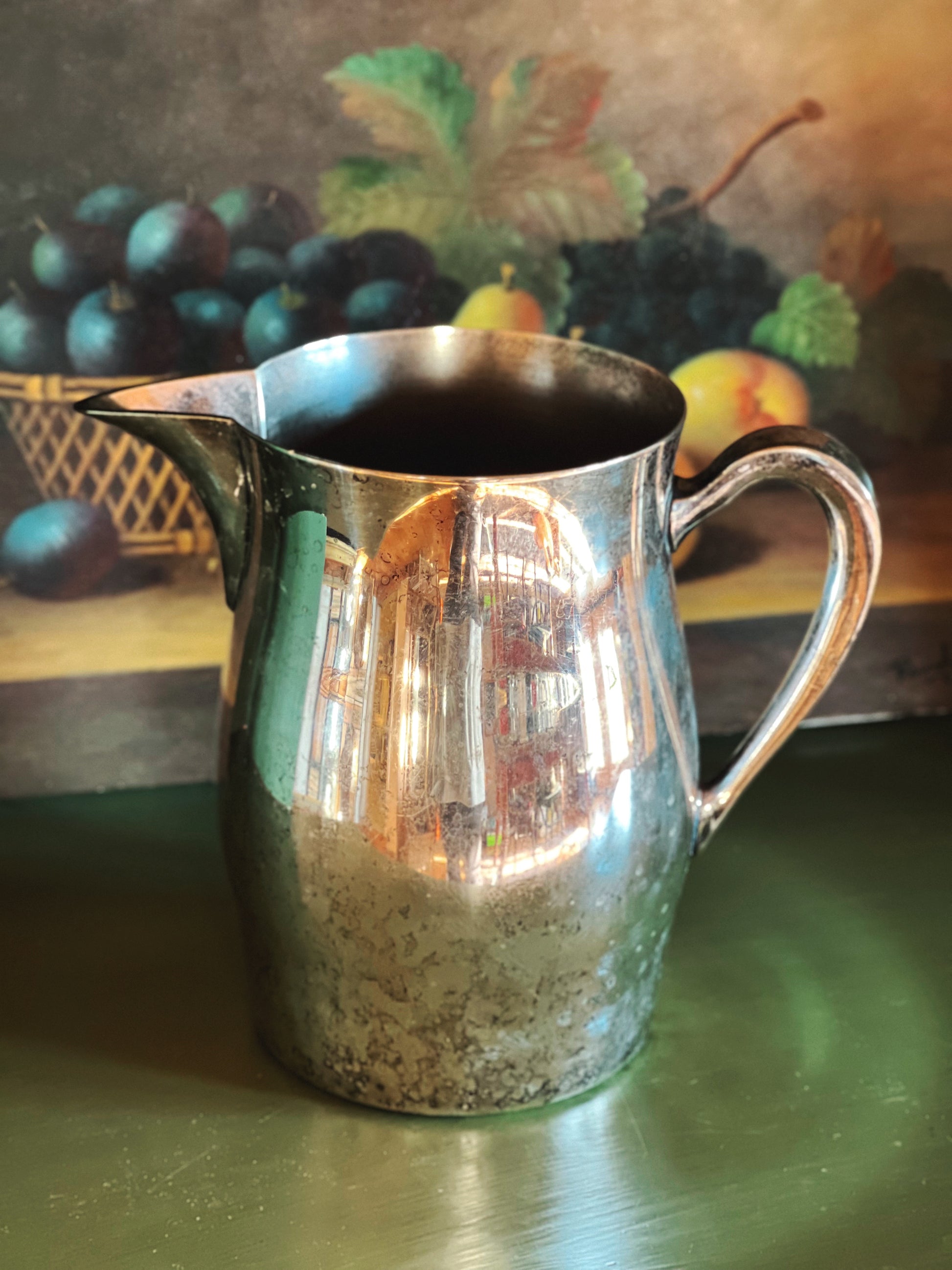 Silver pitcher on a reflective surface with a fruit painting in the background