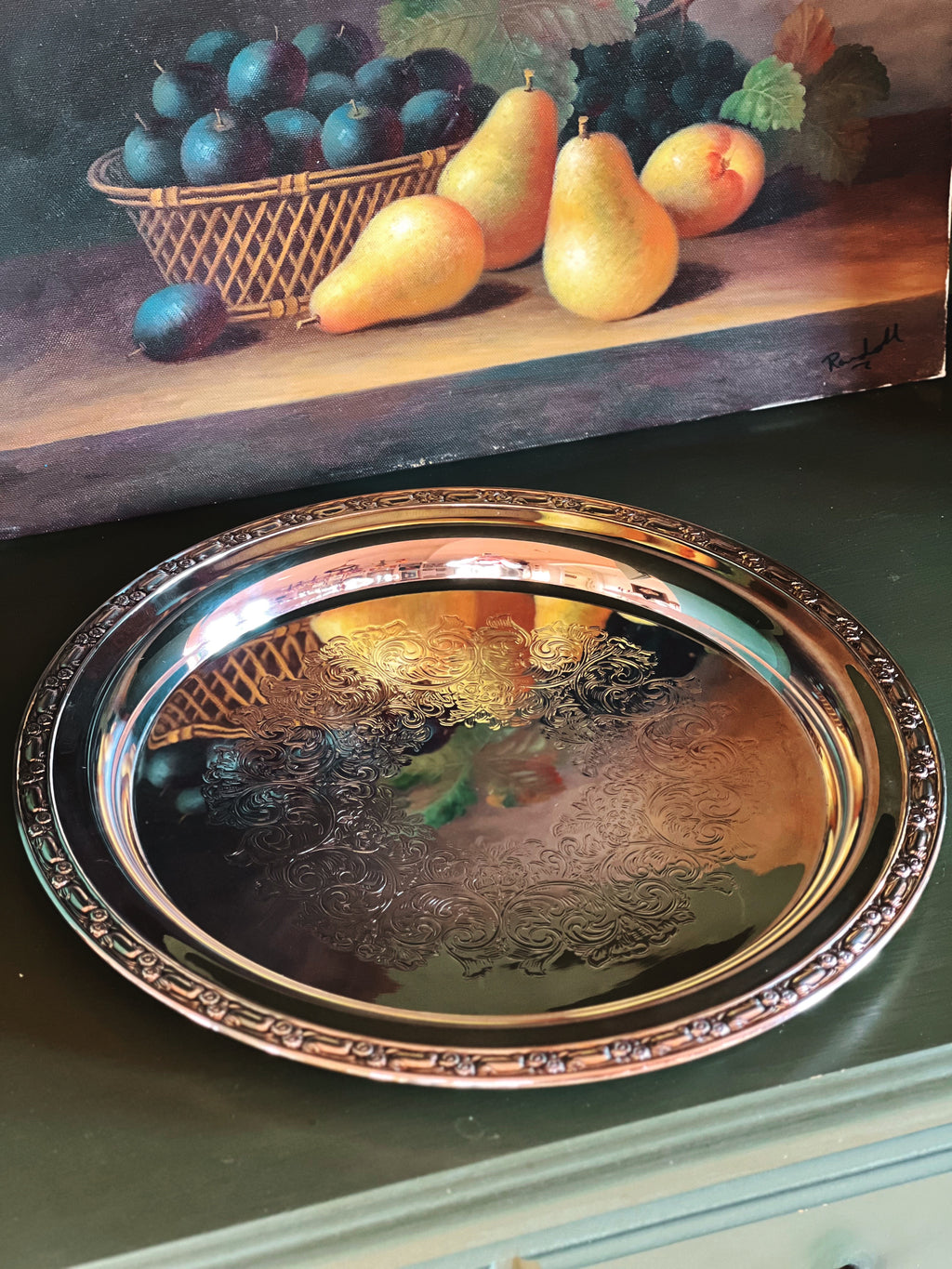 Decorative silver tray with a still life painting of fruit in the background