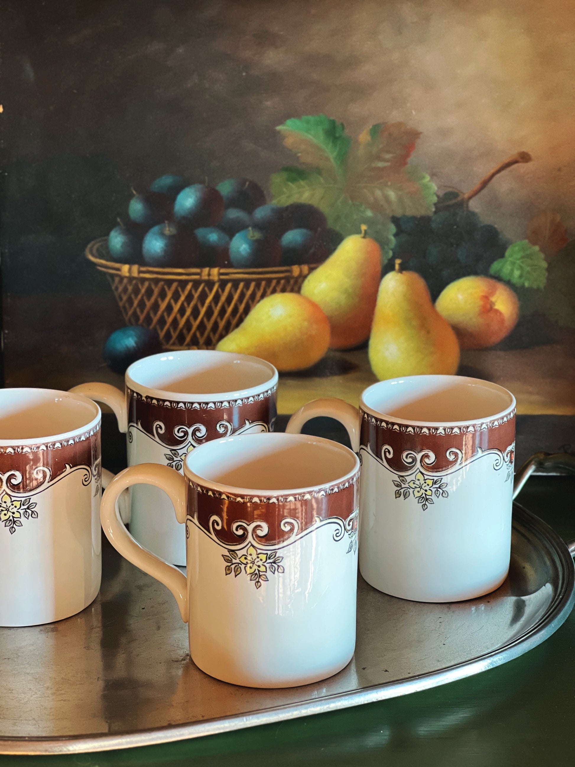 Four ceramic mugs with decorative handles on a tray in front of a still life painting of fruit.