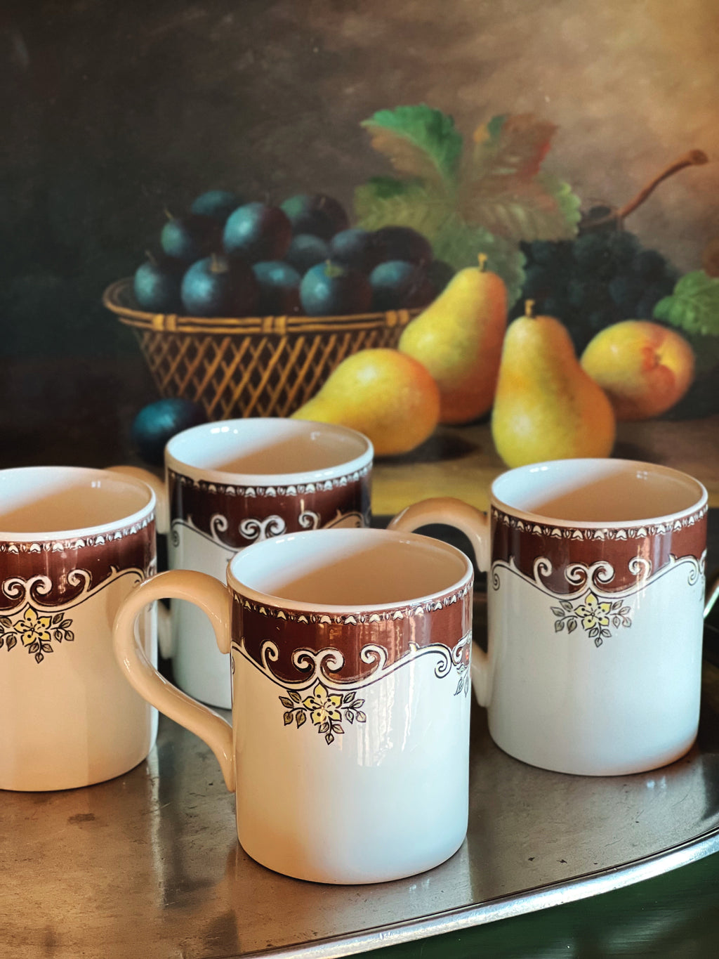 Four mugs with decorative brown rims on a table with a fruit painting in the background.