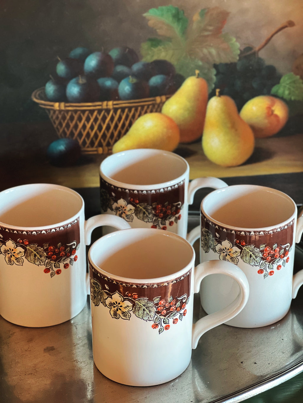 Overhead view of Gibson stoneware mugs with decorative rims on a table in front of a painting of fruit.