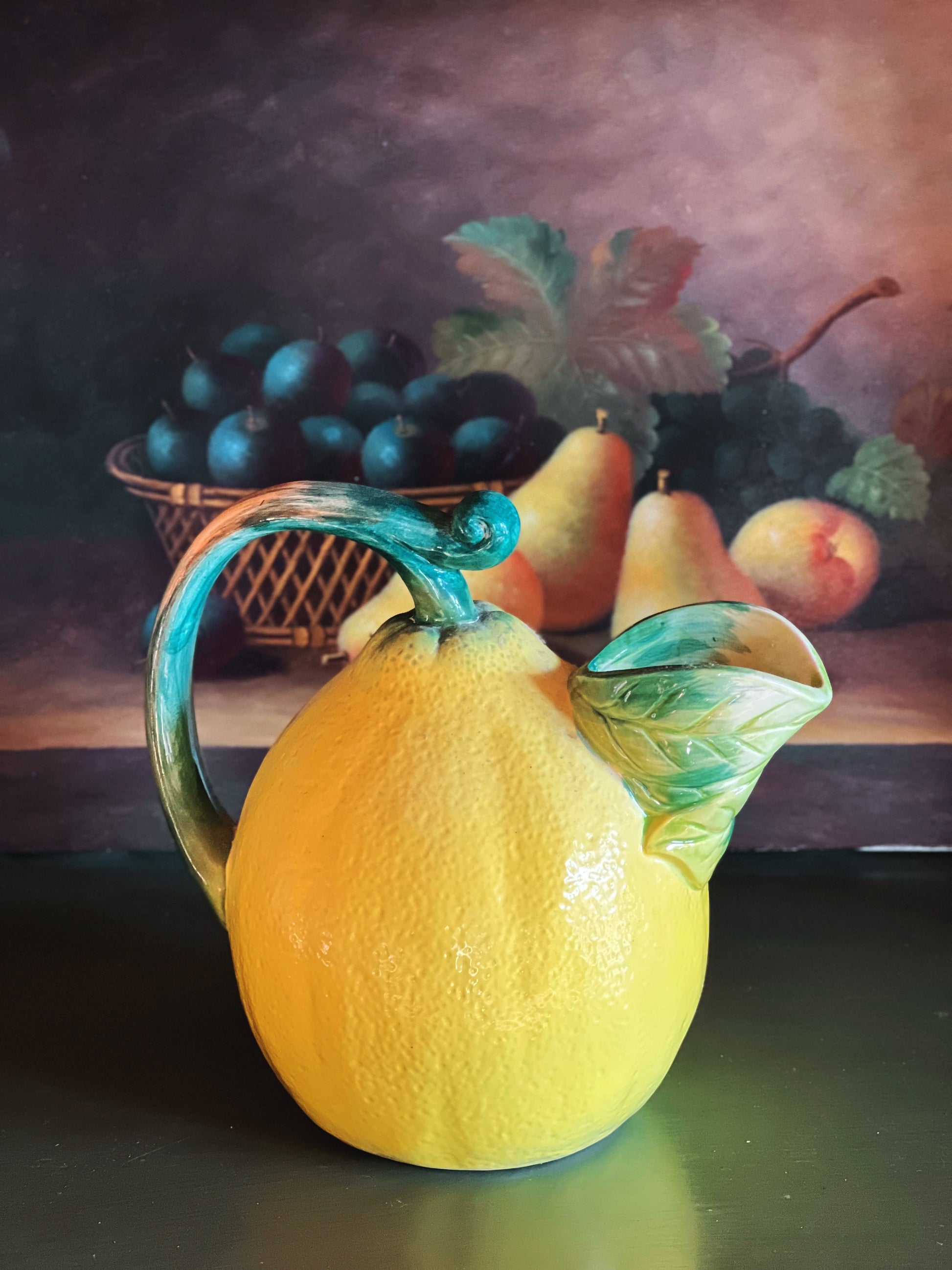 Decorative pitcher shaped like a lemon with a handle and leaf design, set against a still life background.