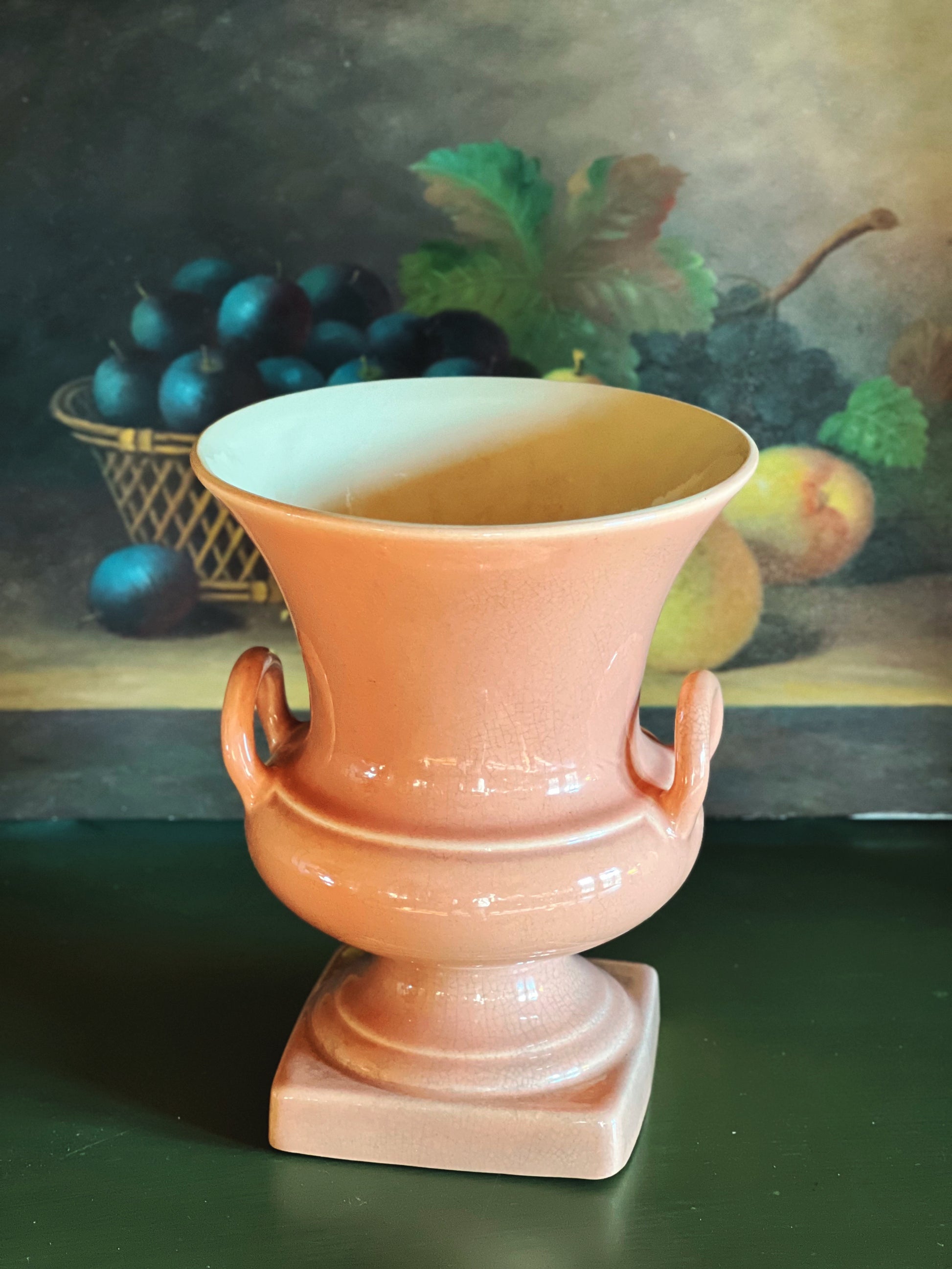 Terracotta-colored urn on a dark surface with a painting of fruit in the background
