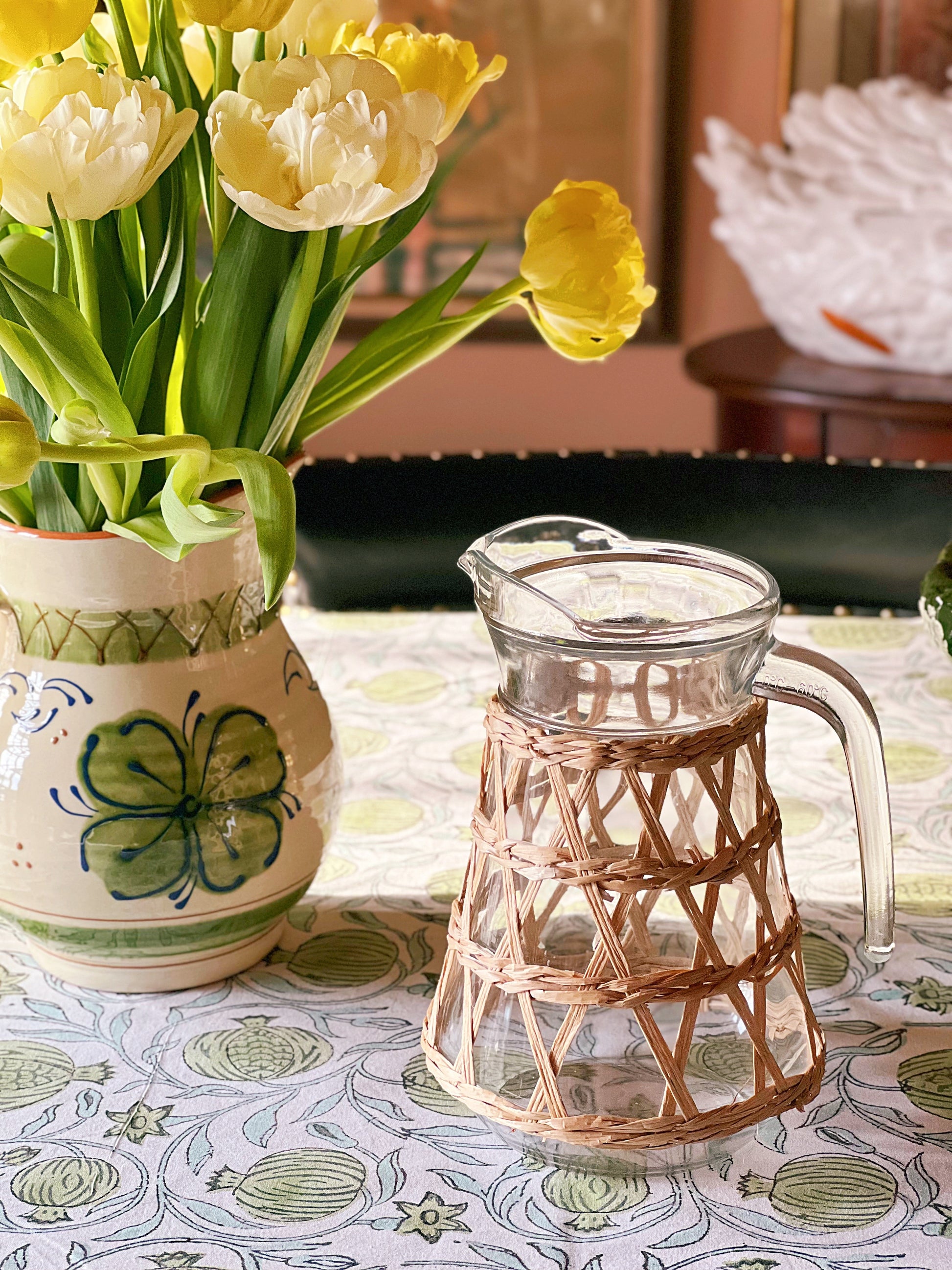 Glass pitcher with woven handle next to a ceramic vase with yellow tulips on a patterned tablecloth.