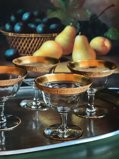 Set of gold-rimmed glass dessert dishes on a reflective surface with fruit in the background.