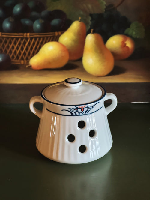 White ceramic pot with blue accents on a dark surface, with pears and a basket in the background.
