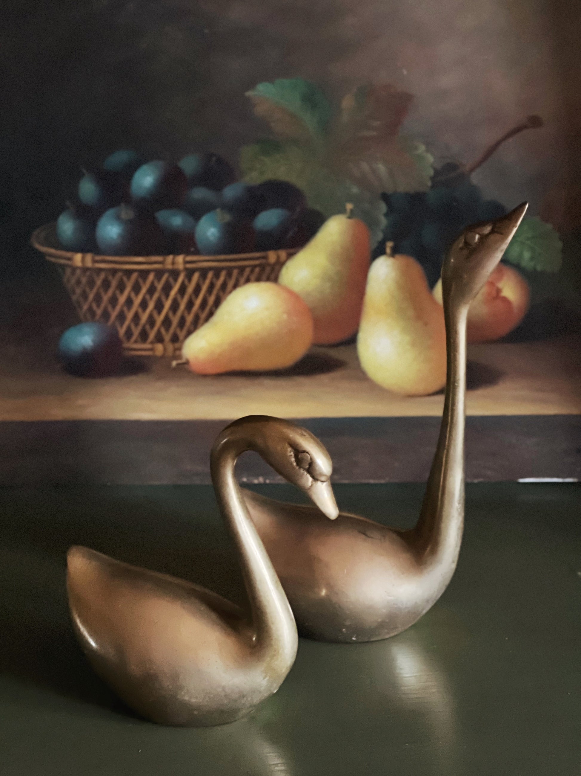 Two bronze swans on a reflective surface with a still life background of fruits and a basket.