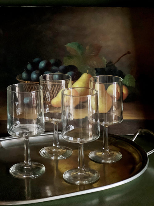Set of four empty wine glasses on a reflective surface with fruit in the background