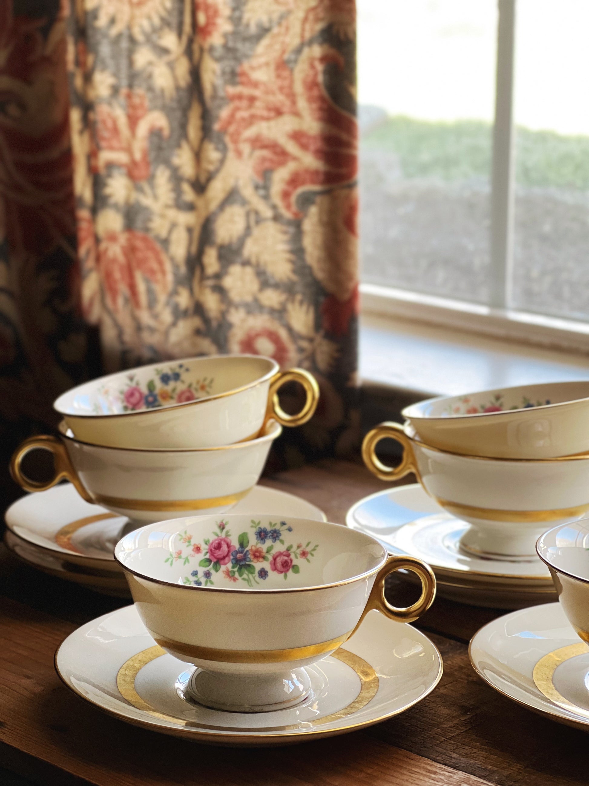 Set of floral teacups and saucers on a wooden surface with a decorative curtain in the background.