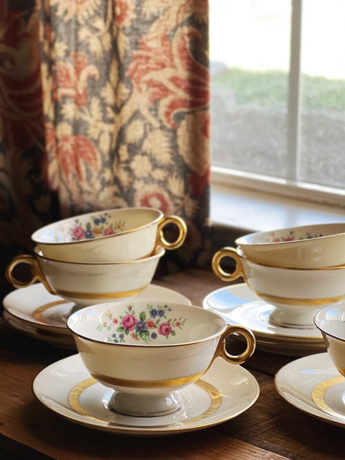 Set of floral teacups and saucers on a wooden surface with a decorative curtain in the background.