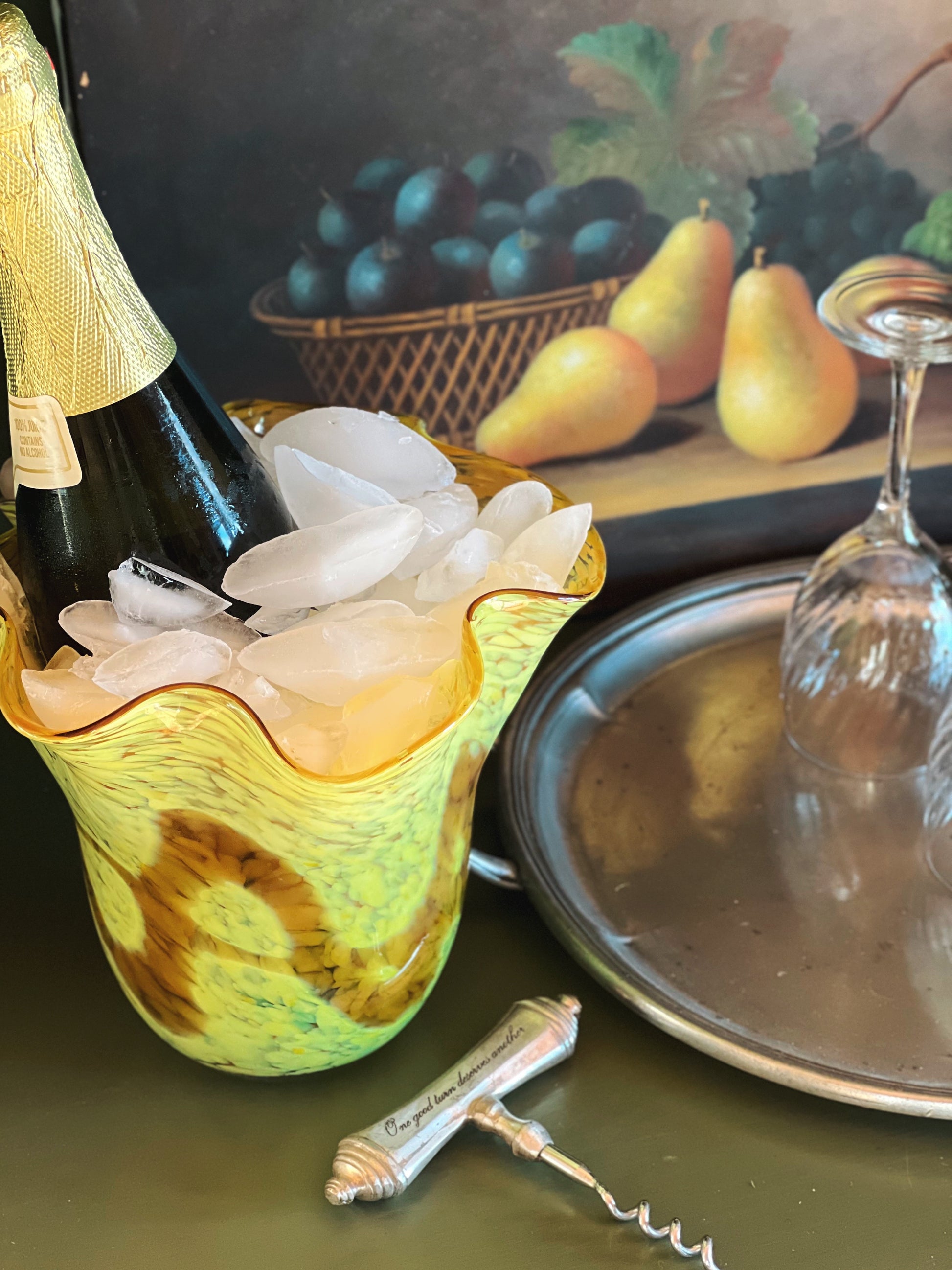 Bottle of champagne with ice in a decorative holder, next to a wine glass and corkscrew on a table.