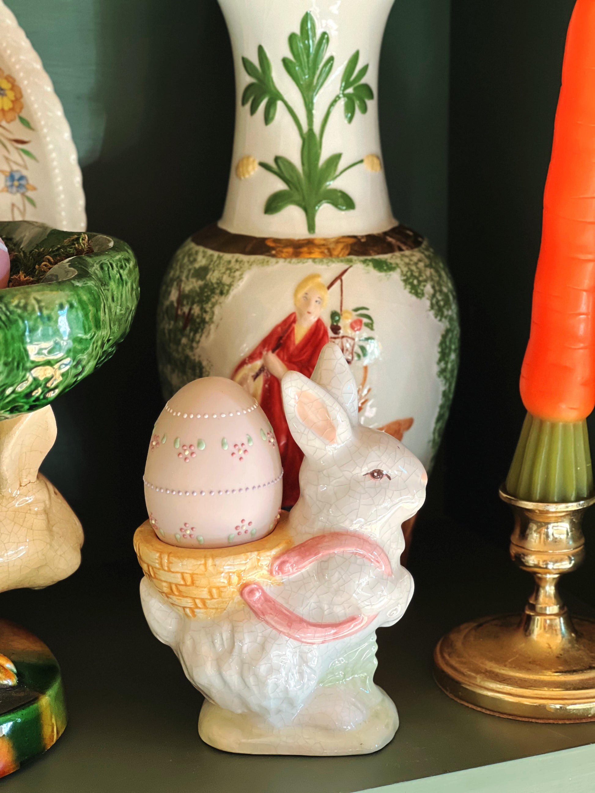 Decorative Easter items including a rabbit figurine with an egg, a vase with a religious scene, and a carrot-shaped object on a dark background.