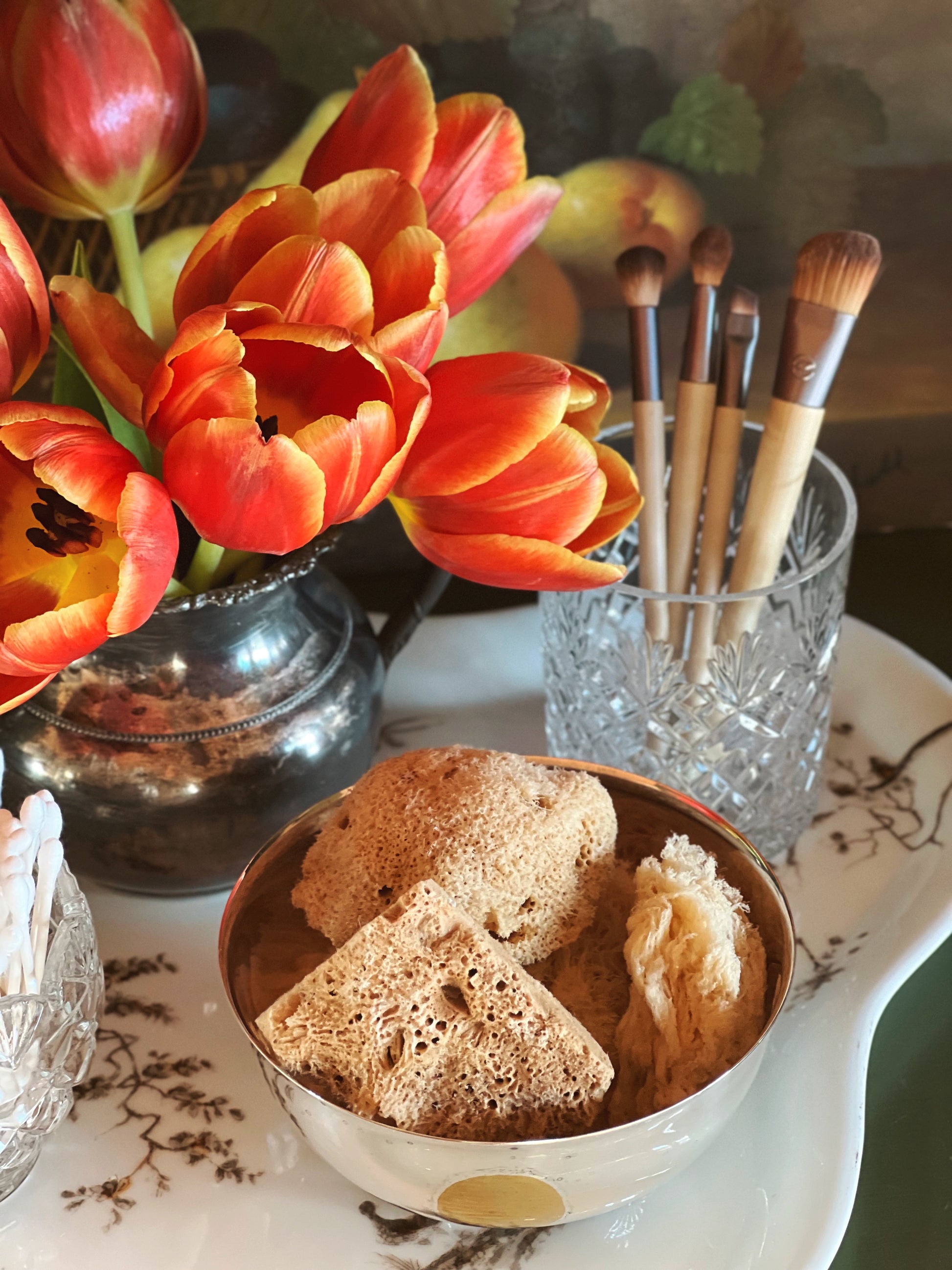 Bowl with sponges, brush holder with makeup brushes, and orange flowers on a decorative table.