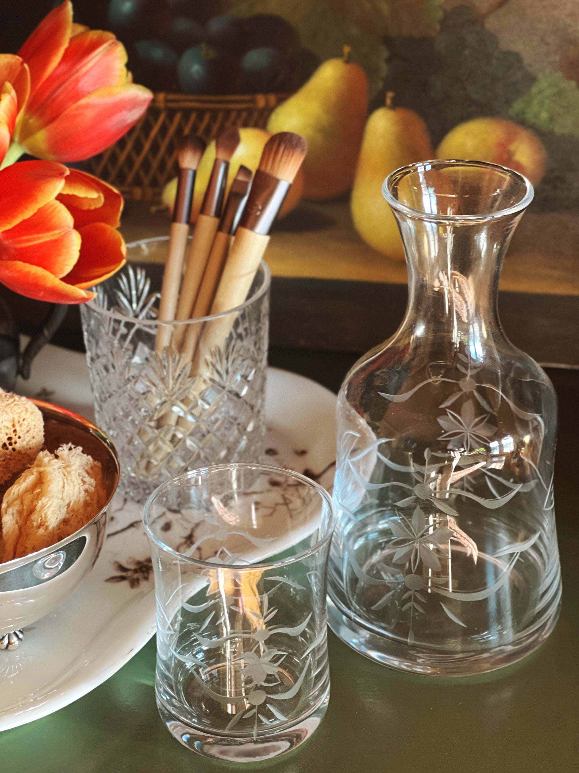 Decorative glassware including a carafe, candle holder, and brush holder on a table with fruit and flowers.