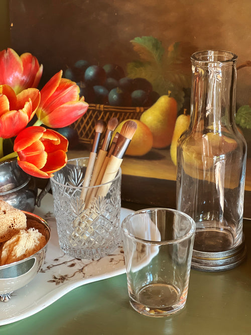 Table setting with makeup brushes in a glass, tulips, and a still life painting in the background.